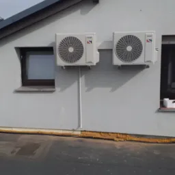 Dwa klimatyzatory marki Sinclair zamontowane na szarej ścianie budynku obok okien, widoczne przewody instalacyjne.