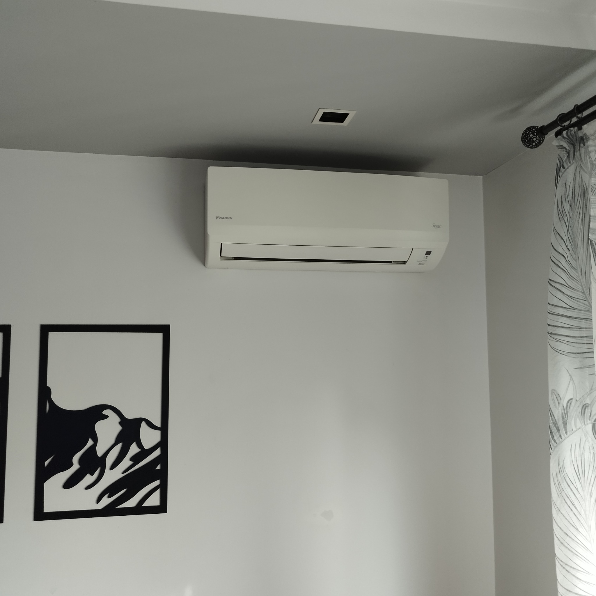 Biały klimatyzator Daikin zamontowany na ścianie w pokoju z nowoczesnym wystrojem, widoczna czarna dekoracja ścienna przedstawiająca górski krajobraz i zasłona w liście palmy.