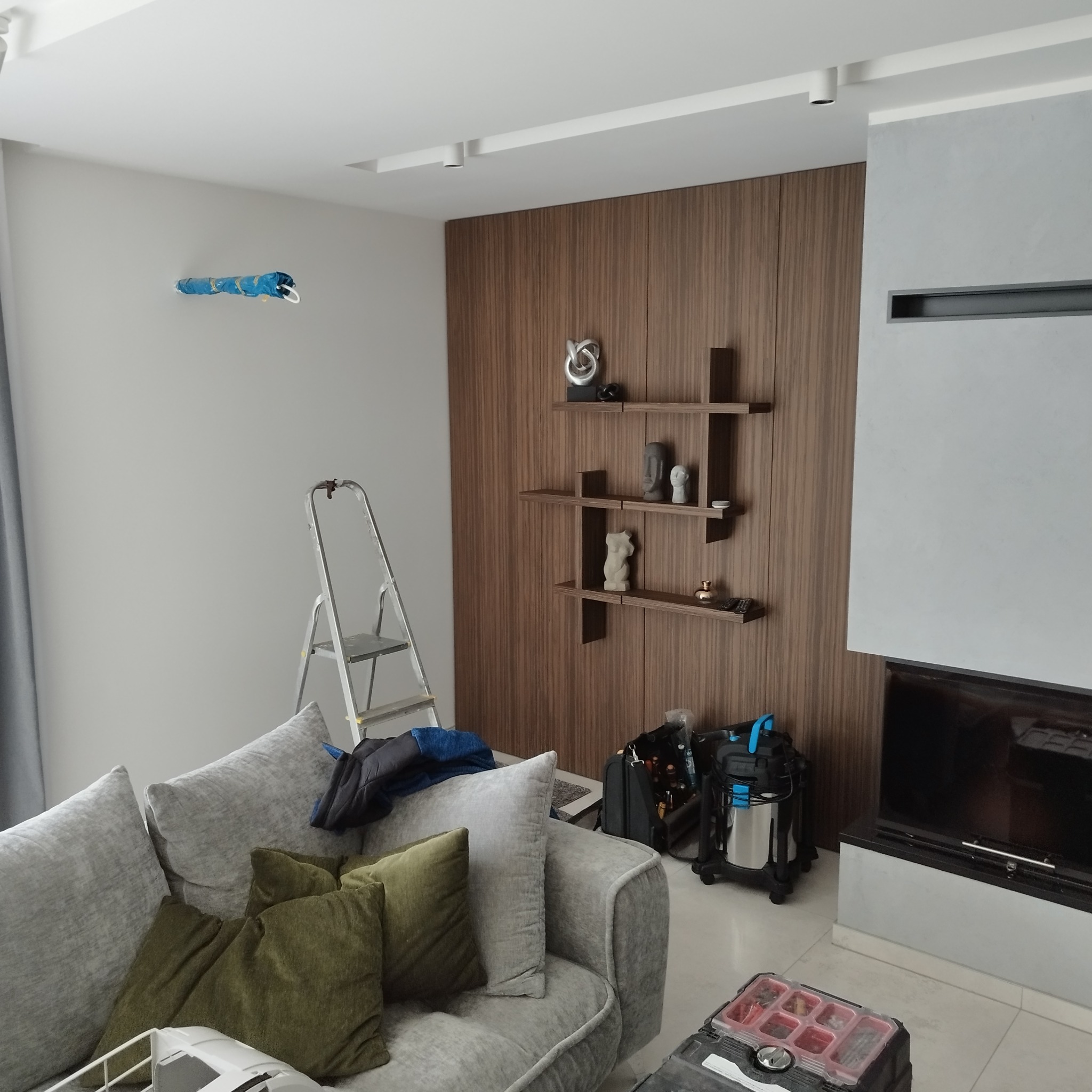 Salon w trakcie remontu: drabina oparta o ścianę, zabezpieczony otwór wentylacyjny, sofa z poduszkami, narzędzia i odkurzacz budowlany przy kominku.