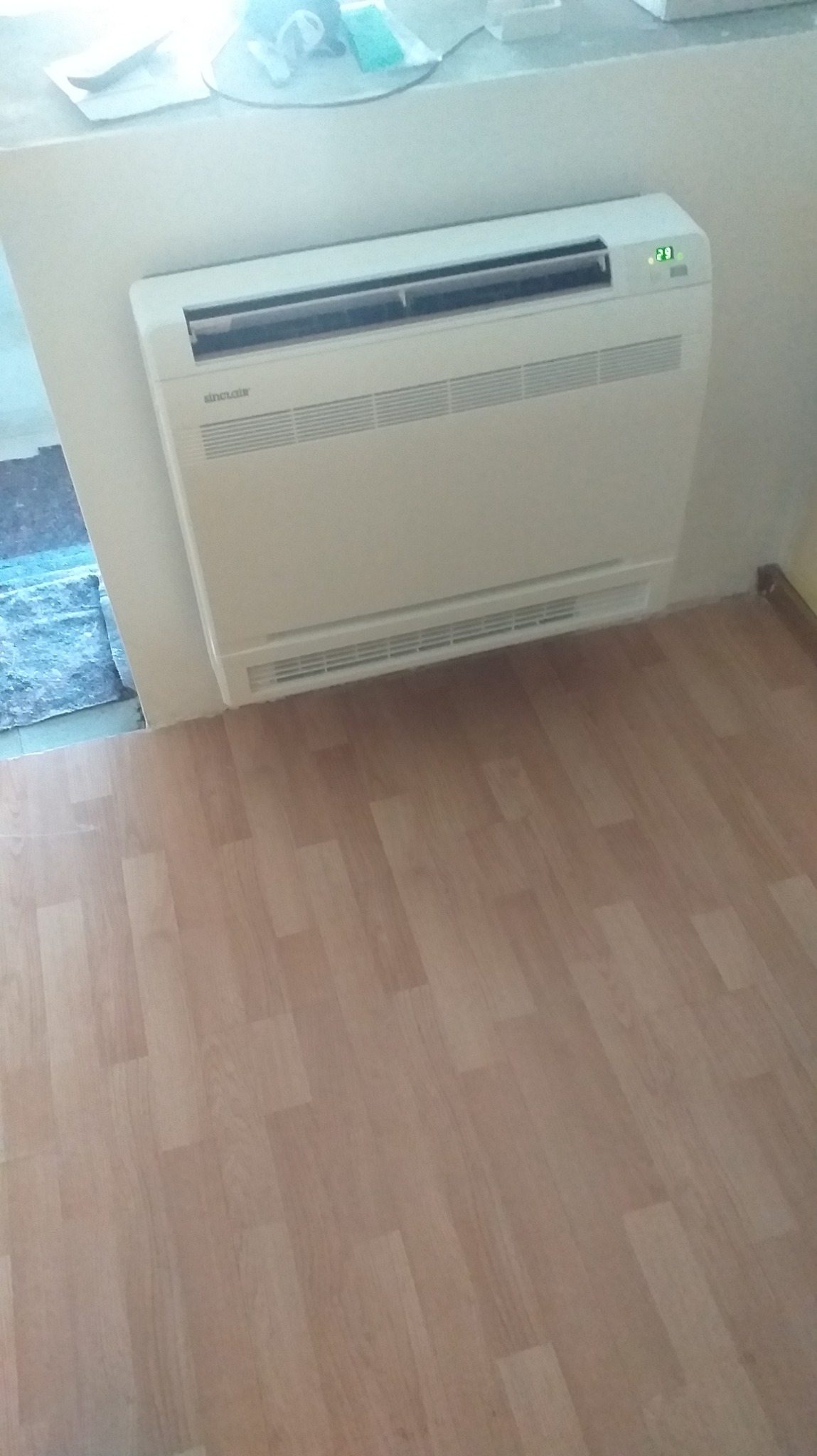 Biały klimatyzator podłogowy marki Sinclair zamontowany przy ścianie, z widocznym wyświetlaczem temperatury 23 stopnie, na tle paneli podłogowych imitujących drewno.