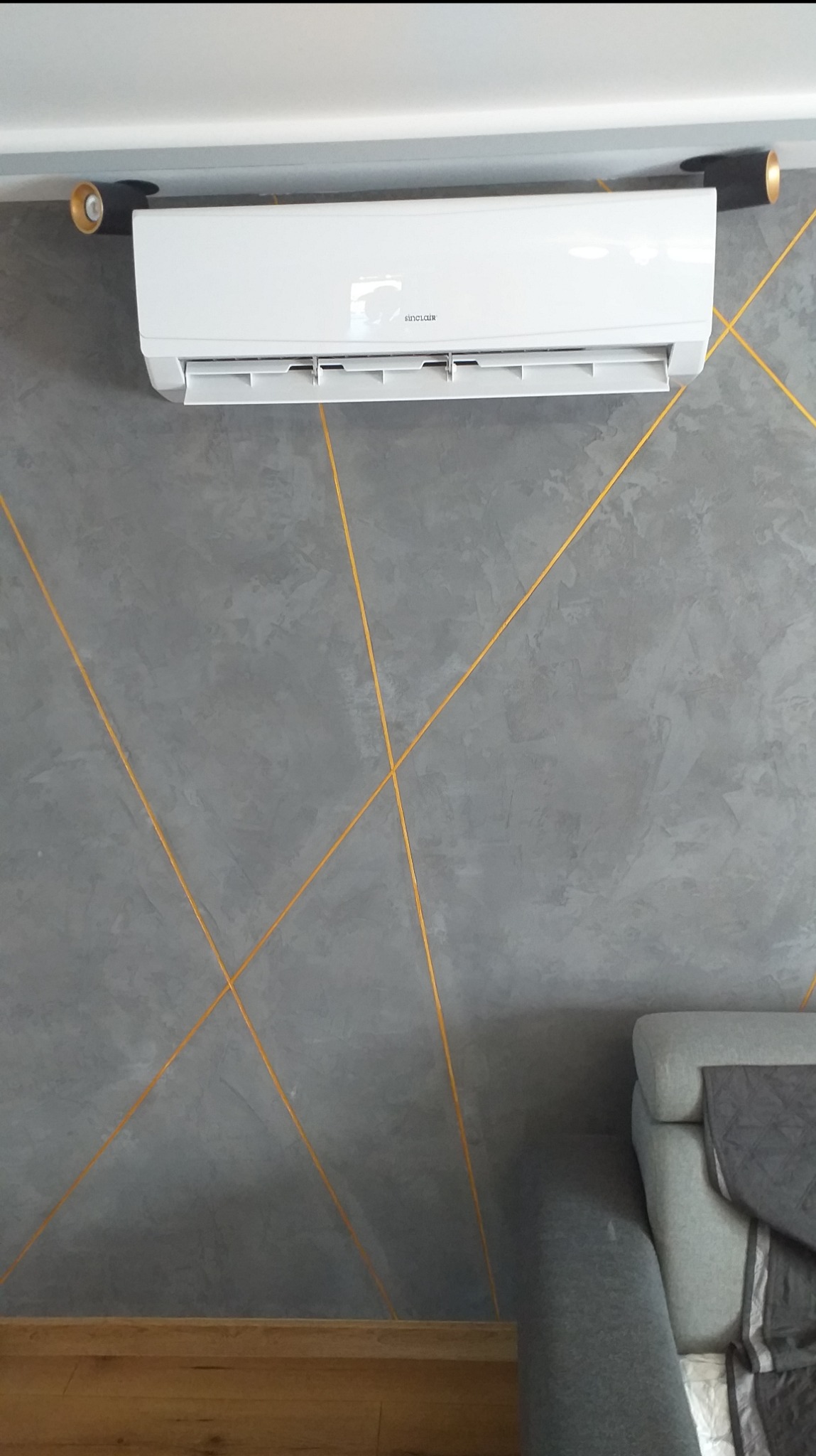 Biała jednostka klimatyzacyjna zamontowana na szarej ścianie z dekoracyjnymi złotymi liniami, widoczne reflektory punktowe i fragment sofy.