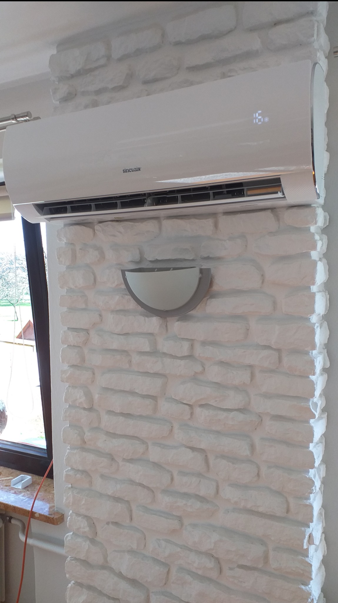 Biały klimatyzator Sinclair zamontowany na ścianie z dekoracyjnej białej cegły, pod nim półokrągła lampa ścienna, widok z okna w tle.