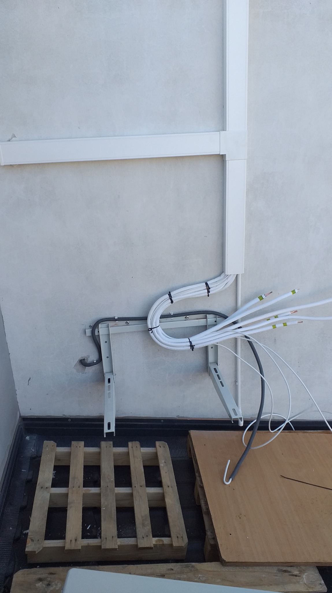 Białe kable zabezpieczone czarnymi opaskami, prowadzone wzdłuż ściany i przymocowane do metalowego wspornika, z widocznymi końcówkami przewodów oraz fragmentem drewnianej palety na podłodze.