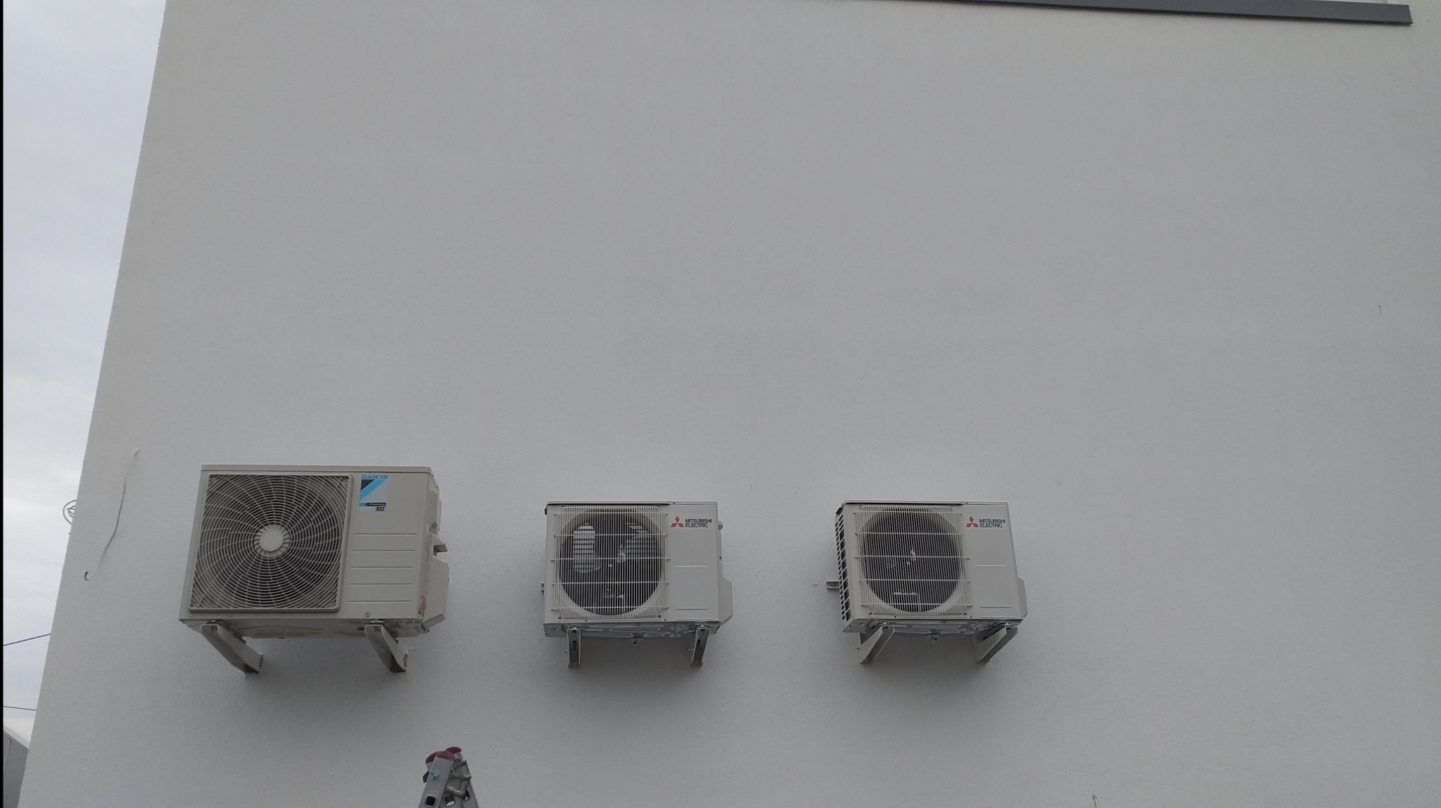 Trzy jednostki zewnętrzne klimatyzacji, Daikin i Mitsubishi Electric, zamontowane na białej ścianie budynku, widoczne fragmenty drabiny, pochmurne niebo w tle.