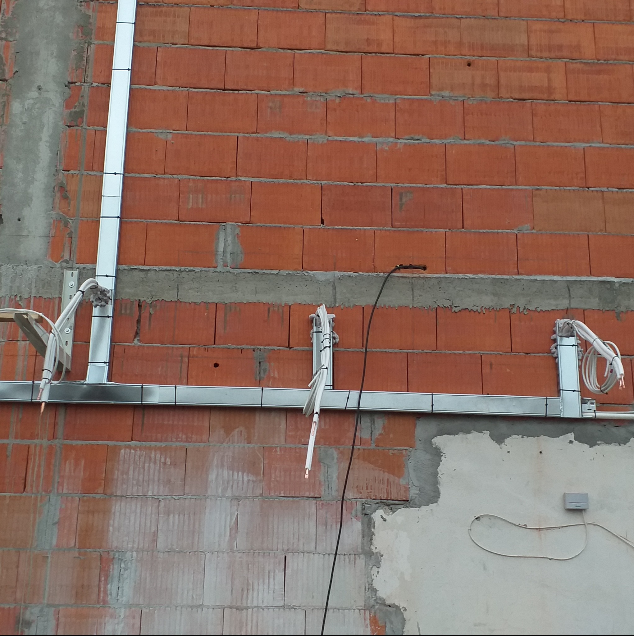 Instalacja elektryczna w korytach i peszlach prowadzona po surowej ścianie z czerwonej cegły dziurawki, widoczne przewody i puszka elektryczna.