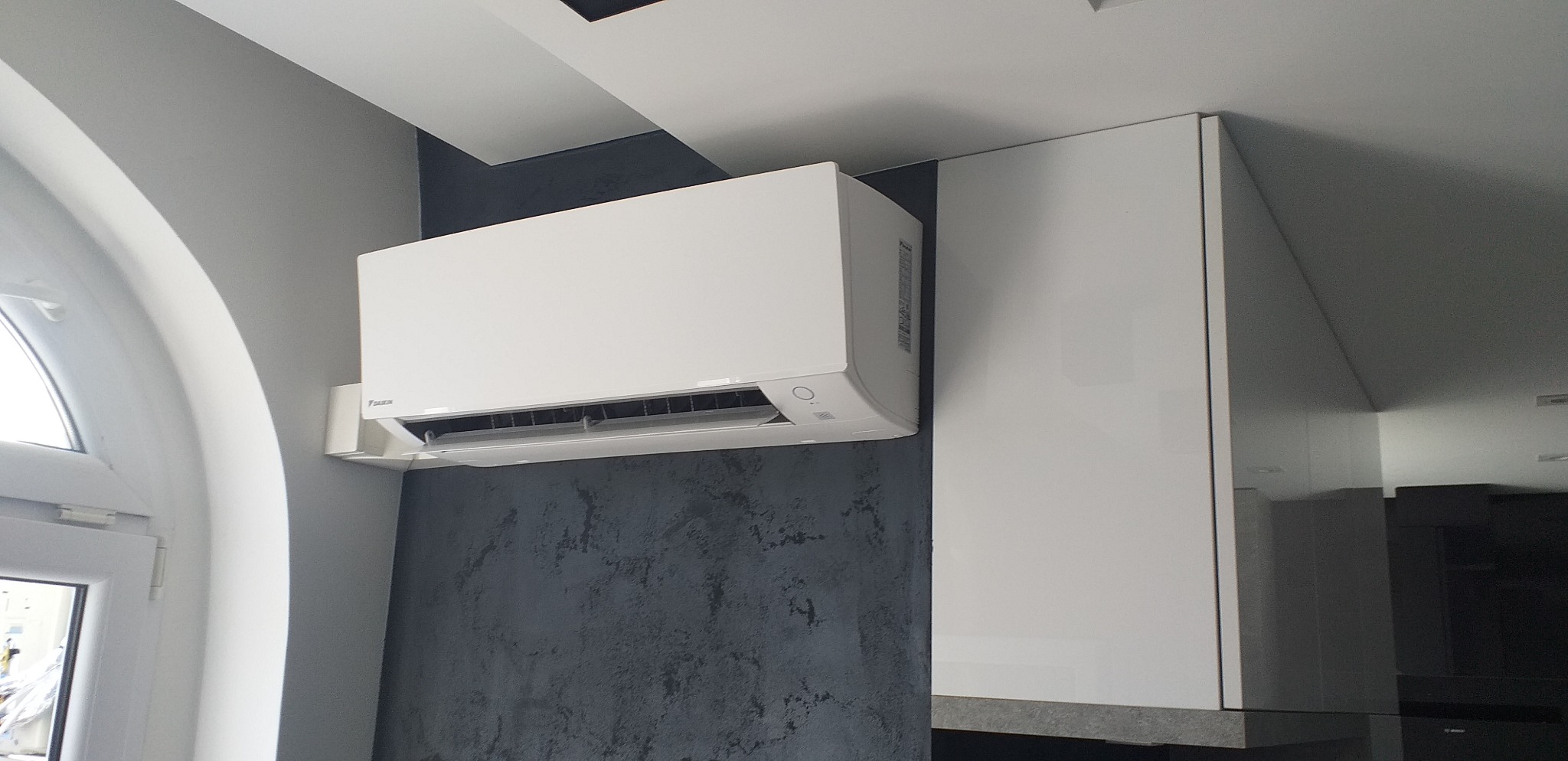 Biały klimatyzator marki Daikin zamontowany na szarej ścianie o fakturze betonu, sąsiadujący z białą zabudową meblową w nowoczesnym wnętrzu.