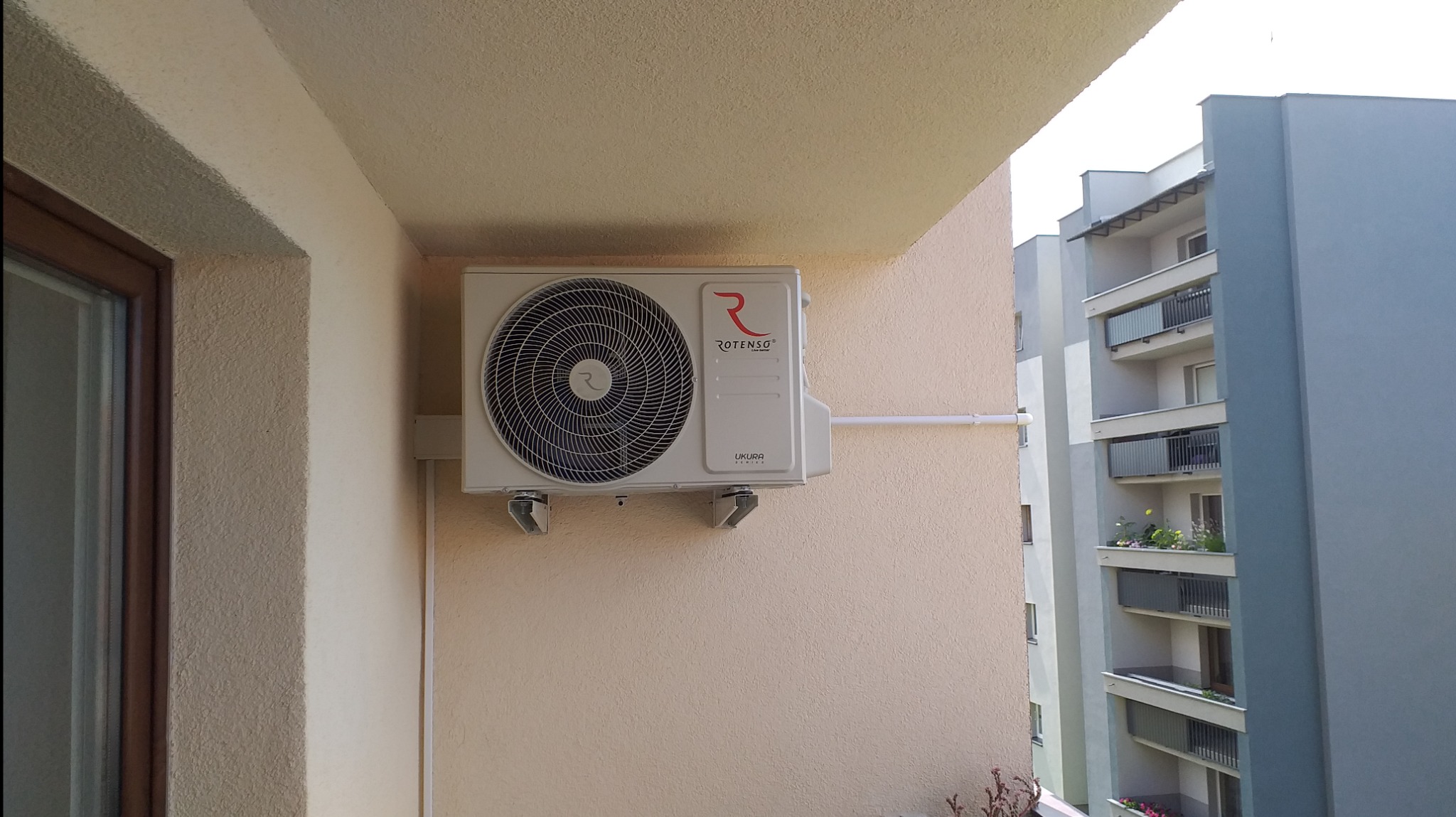 Zewnętrzna jednostka klimatyzatora marki Rotenso zamontowana na balkonie bloku mieszkalnego. Widoczne przewody i uchwyty montażowe.