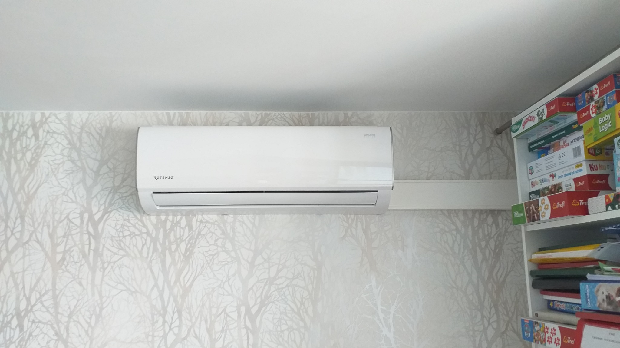Biały klimatyzator Rotenso zamontowany na ścianie z tapetą w delikatny wzór drzew, widoczny fragment regału z grami planszowymi.