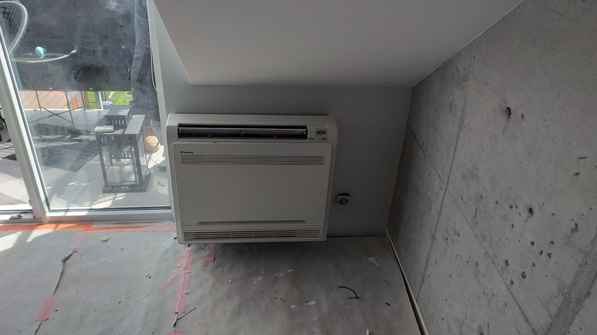 Biała jednostka wewnętrzna klimatyzatora Daikin zamontowana przy ścianie w surowym wnętrzu z widocznymi śladami prac remontowych, zabezpieczona folią podłoga, widok na balkon przez duże okno.