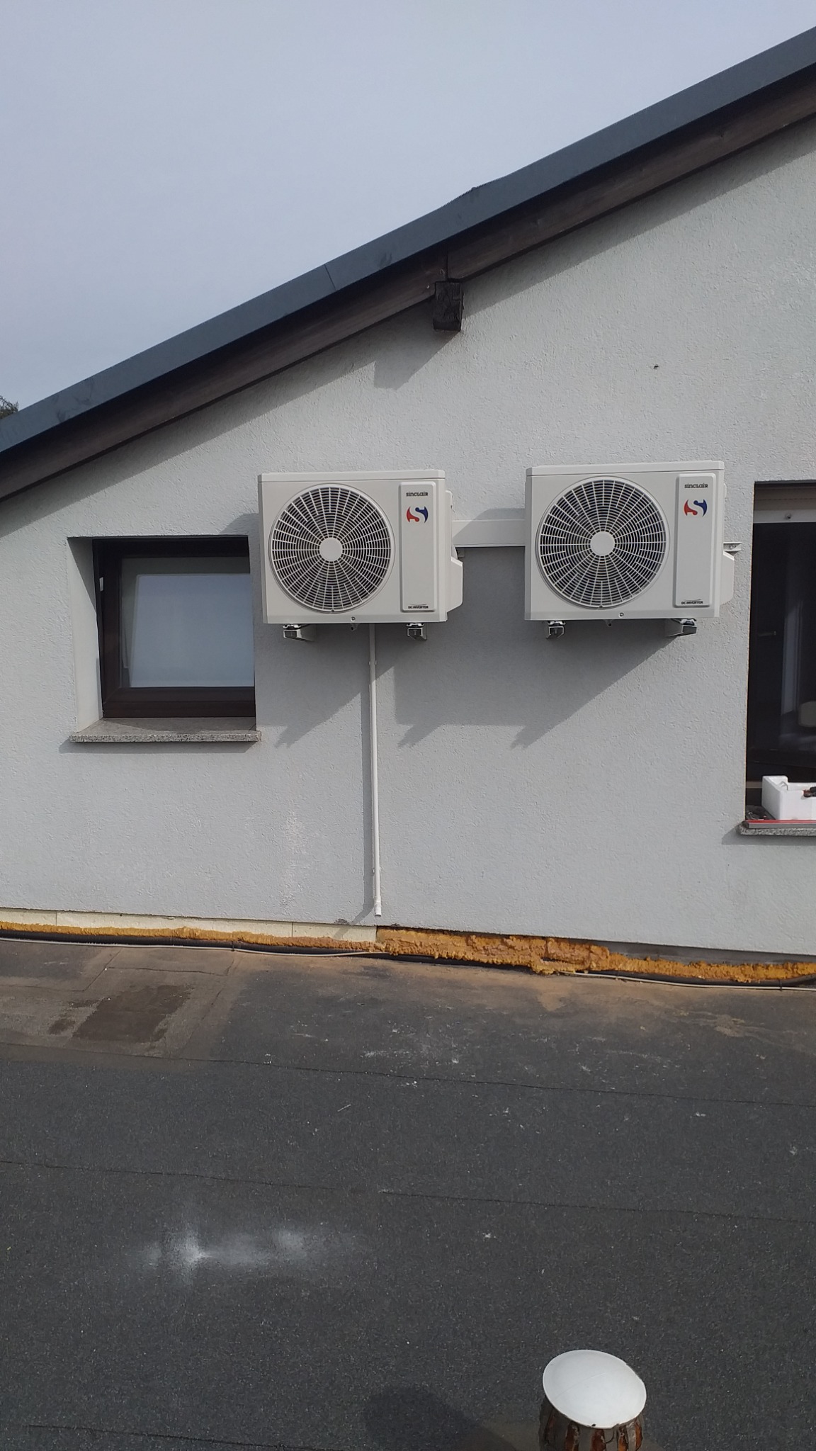 Dwa klimatyzatory marki Sinclair zamontowane na szarej ścianie budynku obok okien, widoczne przewody instalacyjne.