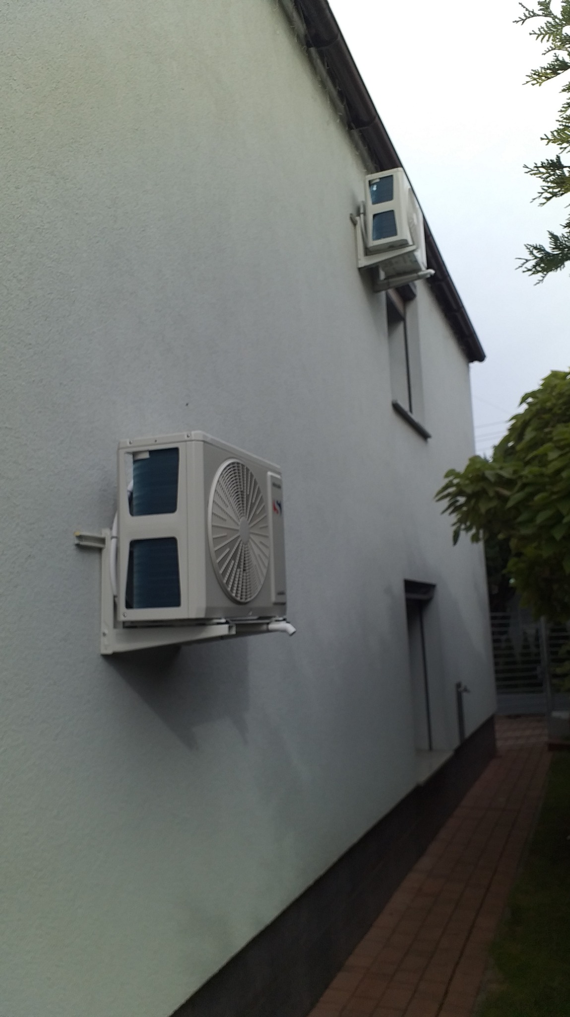 Dwa klimatyzatory zamontowane na zewnętrznej ścianie budynku, widoczne z perspektywy chodnika z kostki brukowej. Większy klimatyzator na niższej kondygnacji, mniejszy nad oknem na wyższej...