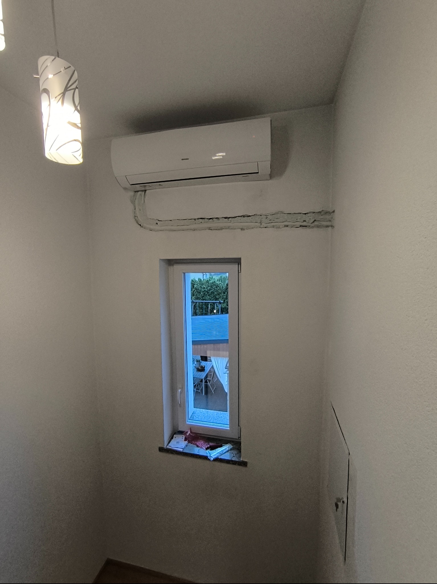 Biały klimatyzator ścienny zamontowany nad oknem, z widocznymi przewodami w listwie maskującej, oświetlony wiszącą lampą.