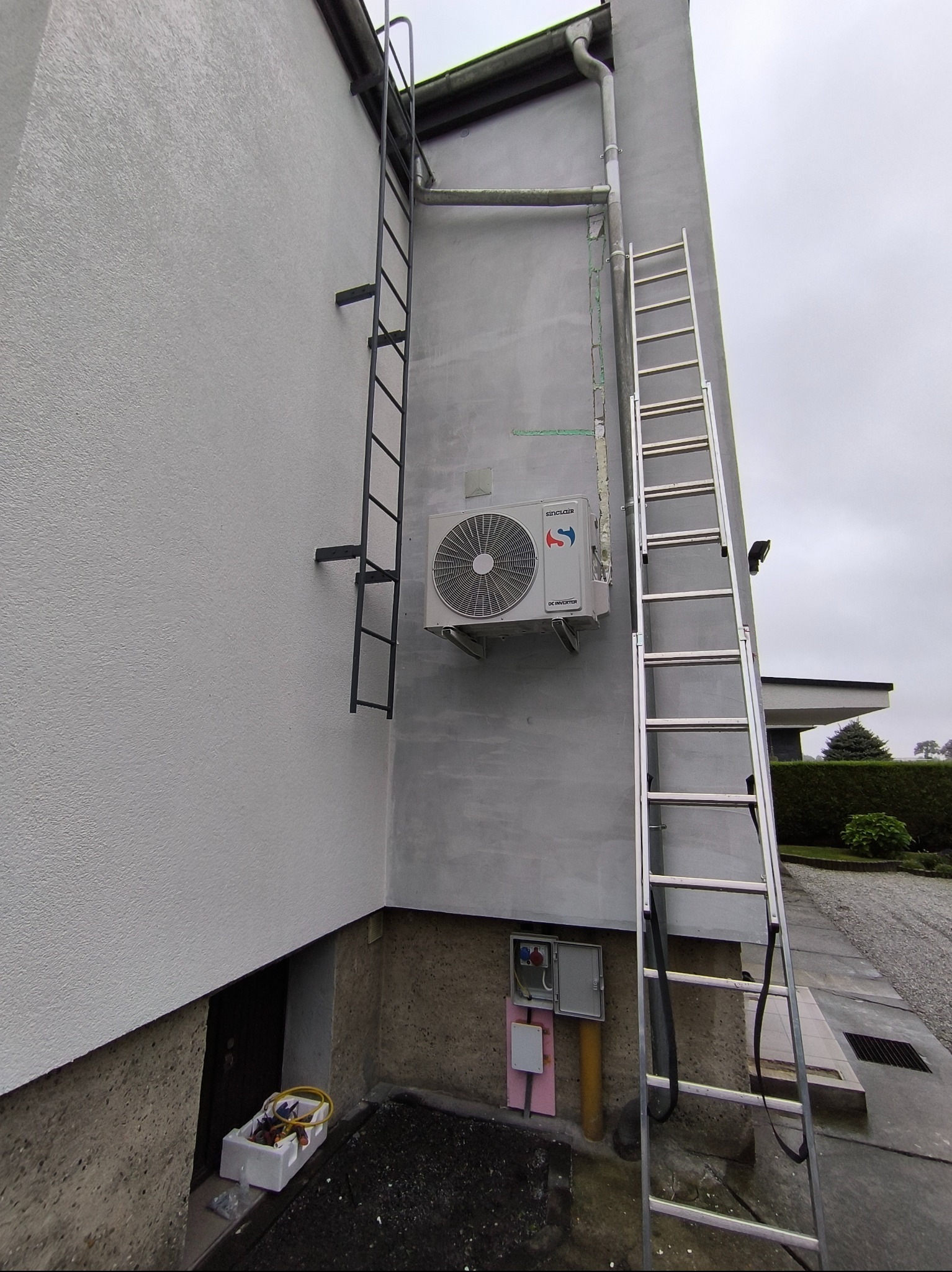 Zewnętrzna jednostka klimatyzacji Sinclair zamontowana na jasnej ścianie budynku, widoczna drabina aluminiowa oparta o ścianę i drabina stalowa zamocowana na stałe, skrzynka elektryczna na dole...
