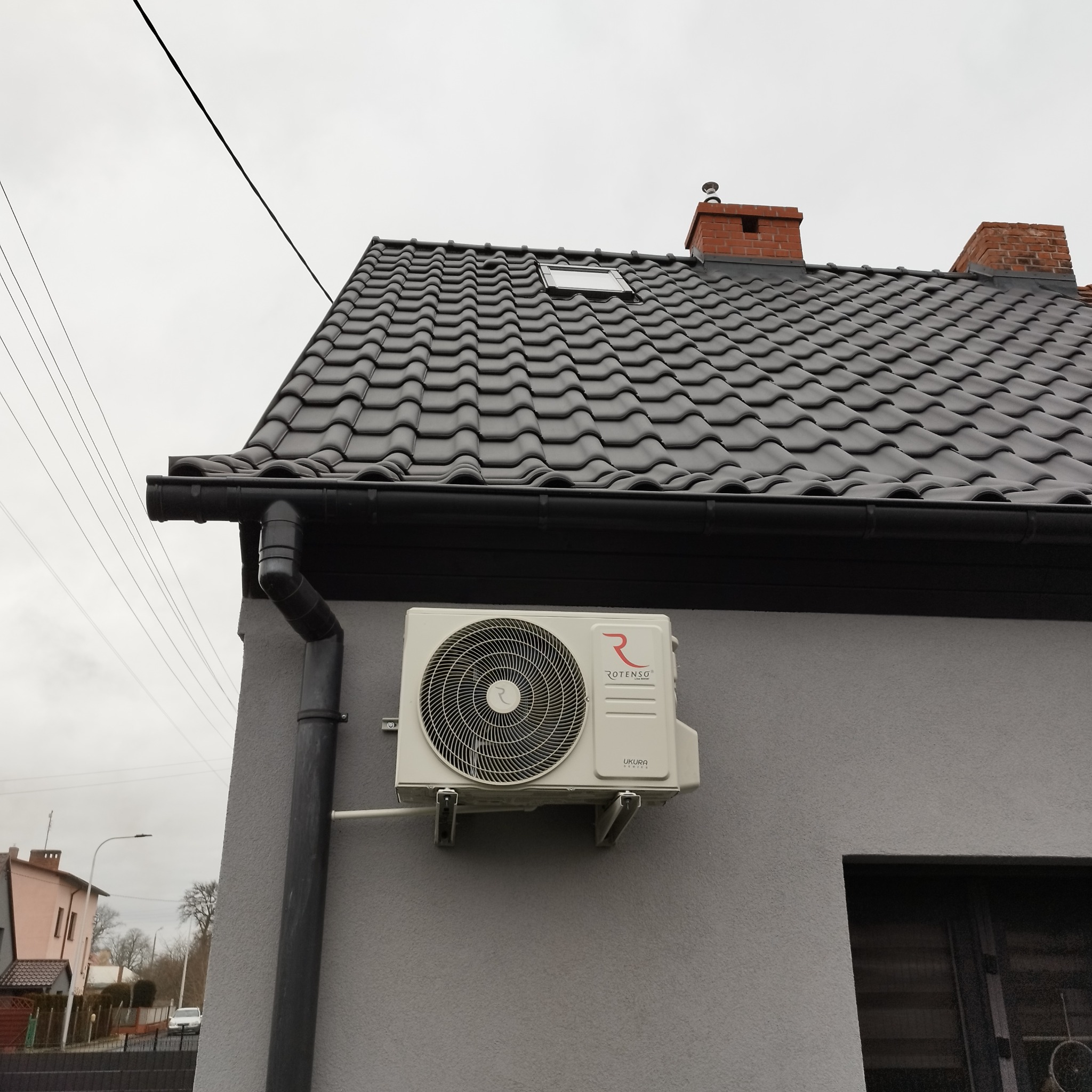 Zewnętrzna jednostka klimatyzatora marki Rotenso zamontowana na szarej ścianie domu z czarną dachówką i orynnowaniem, widok z dołu.