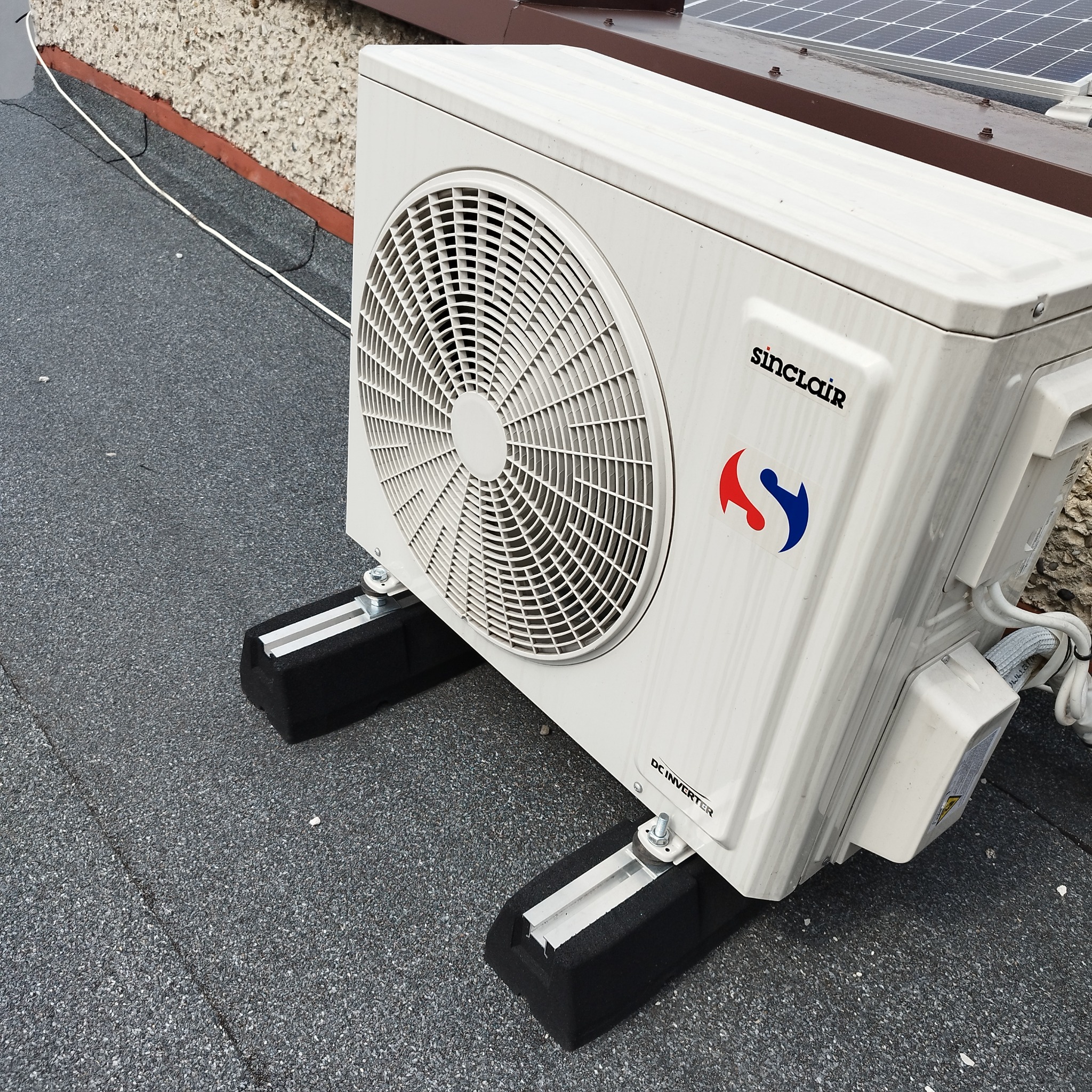 Zewnętrzna jednostka klimatyzatora marki Sinclair, zamontowana na dachu pokrytym papą, z widocznymi panelami słonecznymi w tle.