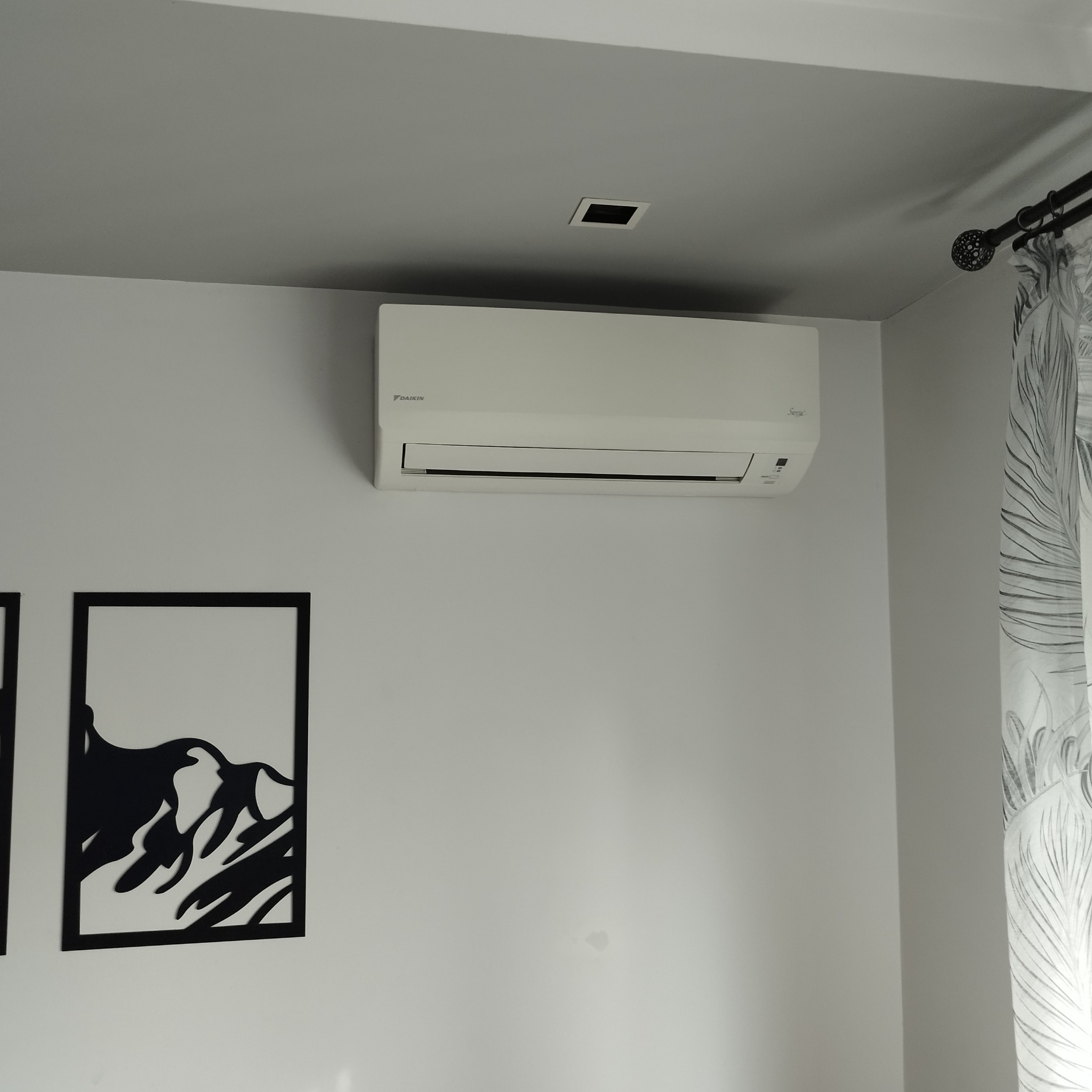 Biały klimatyzator Daikin zamontowany na ścianie w pokoju, obok czarnej dekoracji ściennej z motywem górskim i zasłony w liście.