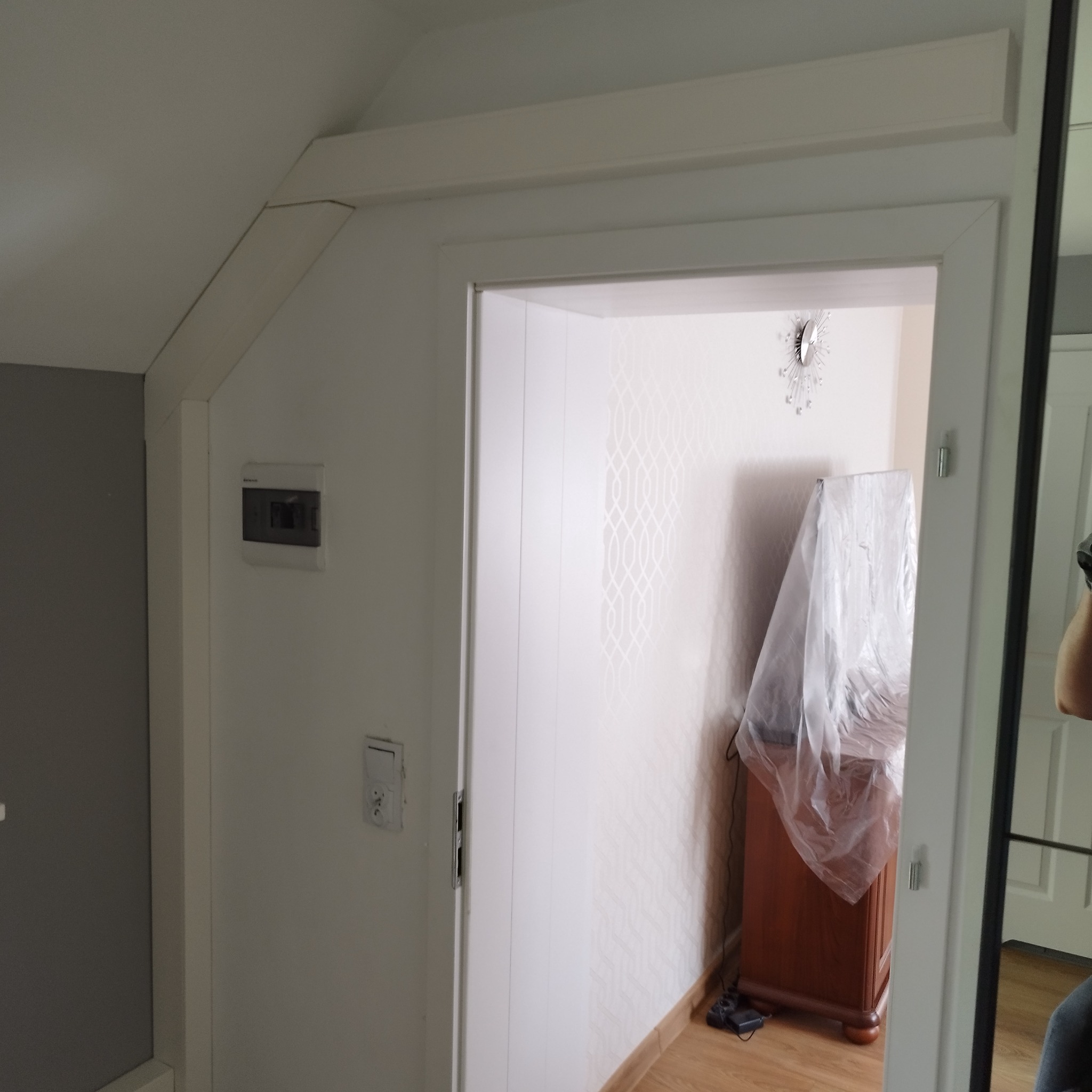 Wnętrze poddasza po remoncie, widoczna ściana z wzorzystą tapetą, skrzynka elektryczna na ścianie, ościeżnica drzwiowa i mebel zabezpieczony folią malarską.