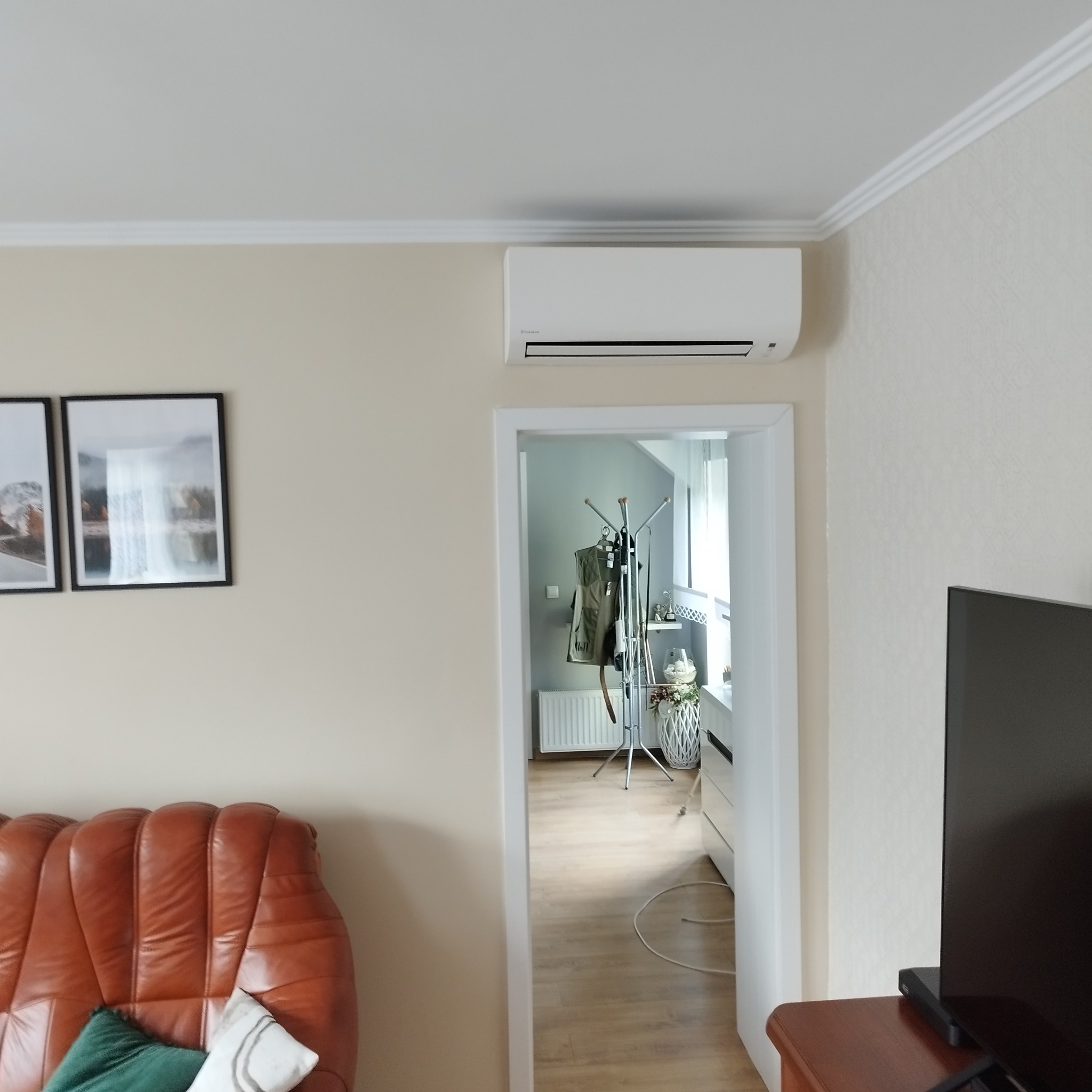 Biały klimatyzator ścienny Daikin zamontowany w jasnym salonie nad drzwiami, widok z boku na skórzaną kanapę i telewizor, w tle widoczny pokój z wieszakiem na ubrania.