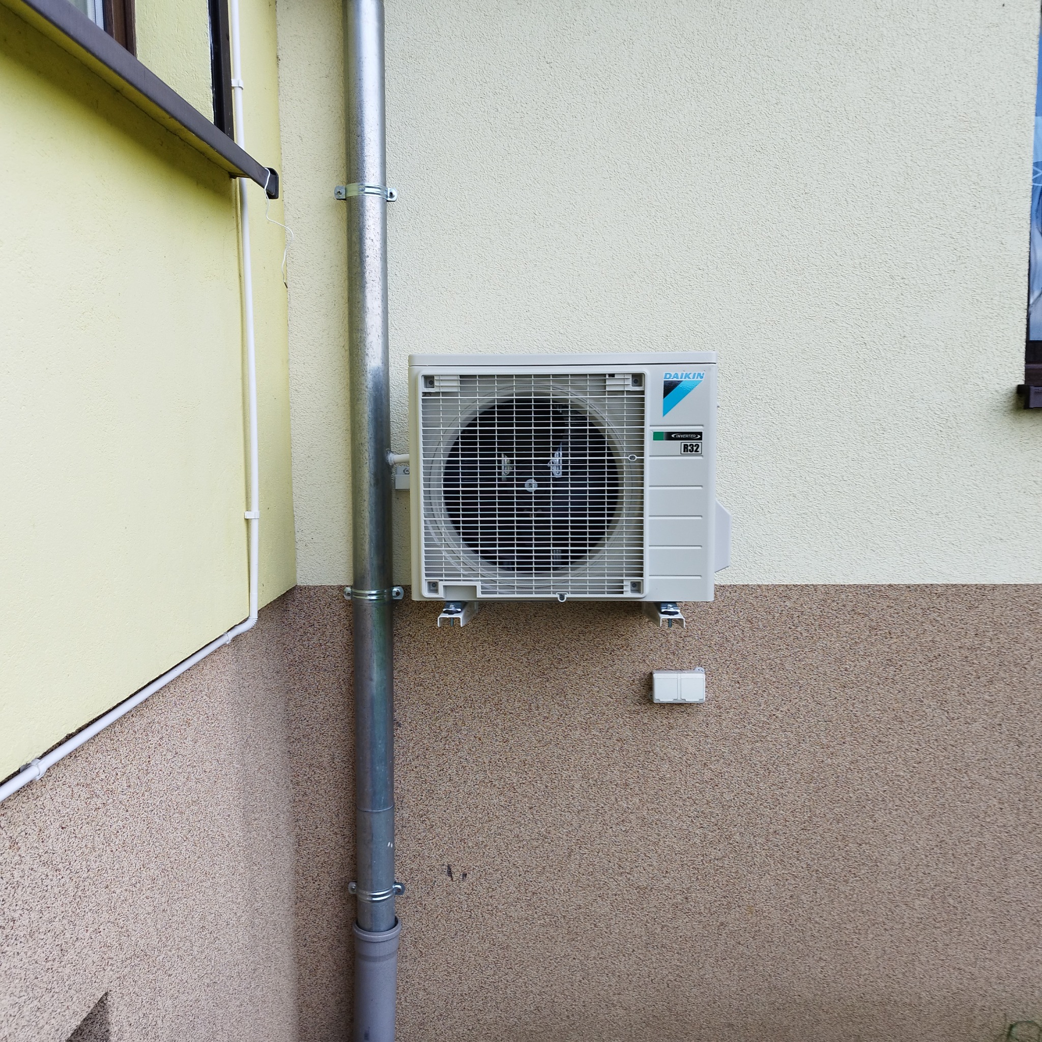 Zewnętrzna jednostka klimatyzatora Daikin zamontowana na ścianie budynku z rurą spustową obok