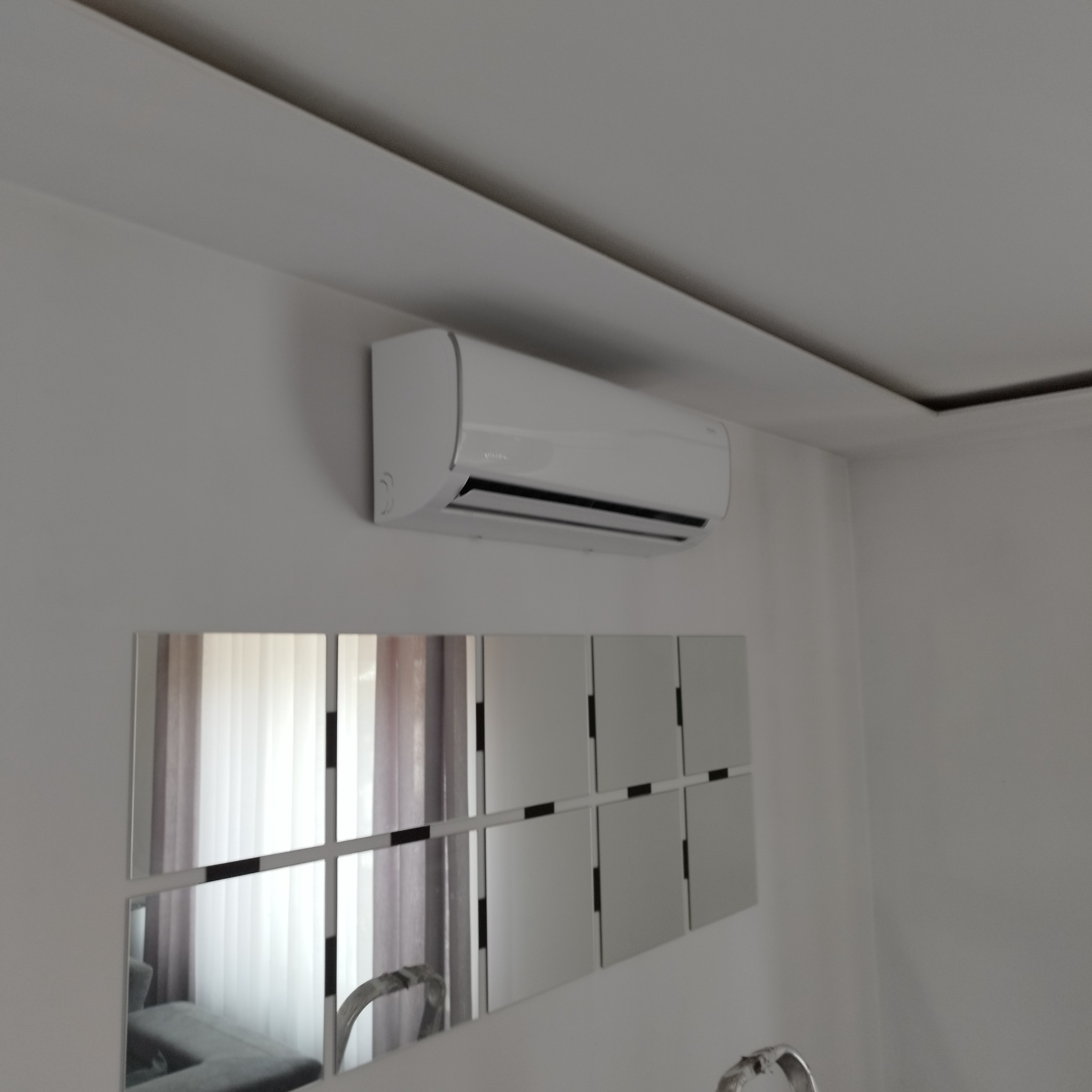 Biały klimatyzator zamontowany na ścianie nad dekoracyjnym panelem z luster, widoczny fragment drabiny.