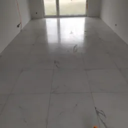 Ułożone, duże, kwadratowe płytki podłogowe imitujące marmur w pustym, jasnym pomieszczeniu z oknem balkonowym zakrytym folią. Na podłodze leży poziomica.