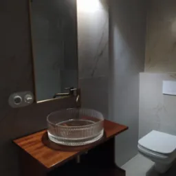 Wykończona łazienka z marmurowymi płytkami, wiszącą toaletą i umywalką z przezroczystego szkła na drewnianej szafce, oświetlona górnym światłem.