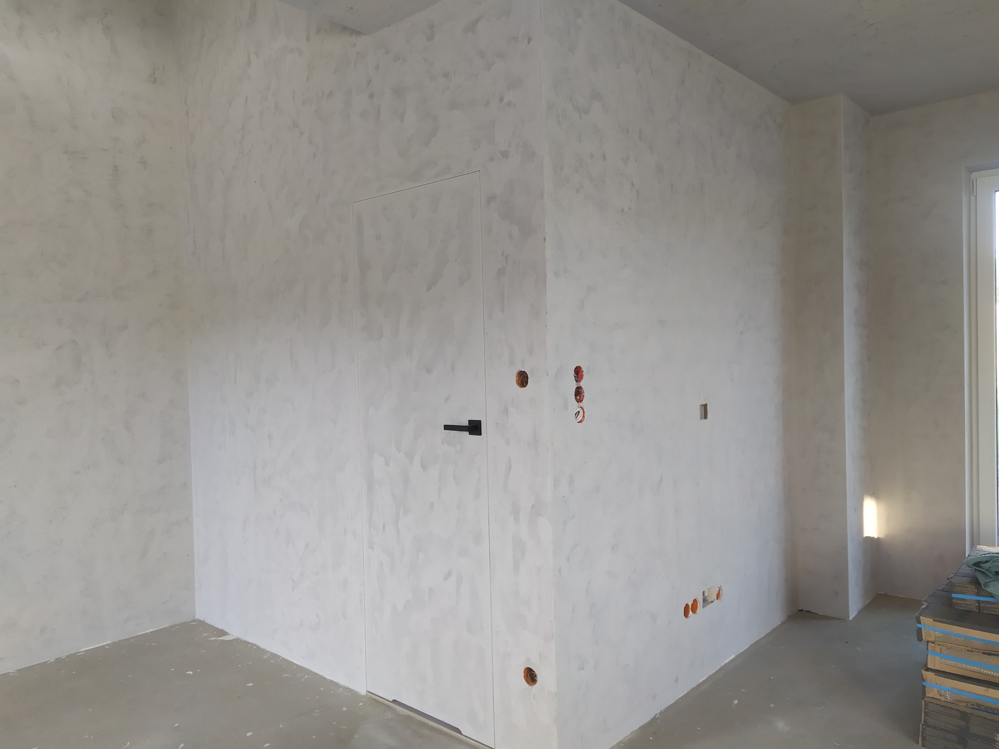 Wnętrze wykończonego pokoju z dekoracyjną, jasną ścianą o fakturze chmur, białe drzwi z czarną klamką, widoczne gniazdka elektryczne i przygotowane podłoże pod panele.