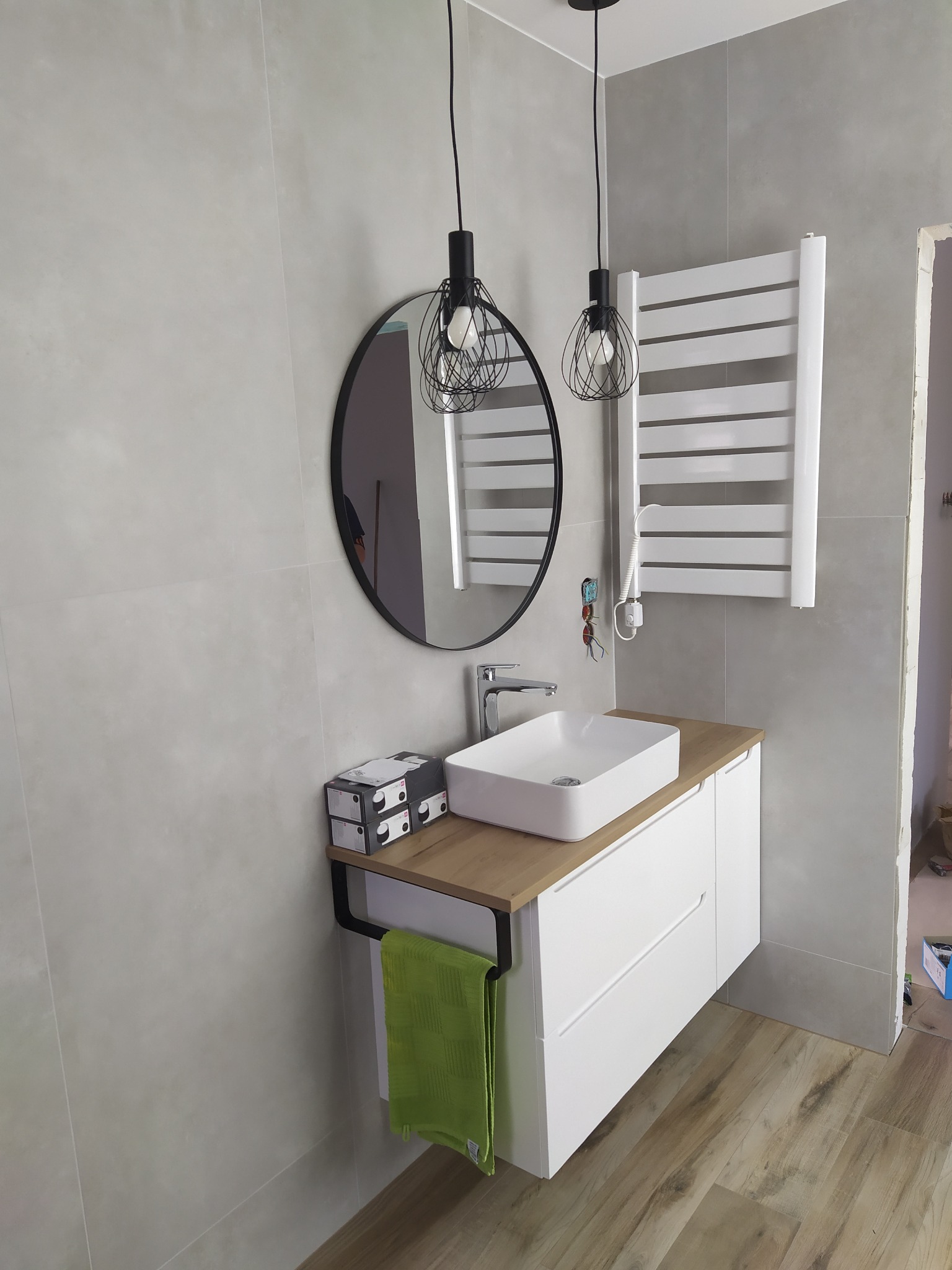 Nowoczesna łazienka z umywalką nablatową, okrągłym lustrem, grzejnikiem drabinkowym i wiszącymi lampami z metalowymi kloszami, wykończona płytkami w odcieniach szarości i drewnopodobną podłogą.