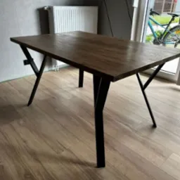 Stół z blatem z ciemnego drewna i czarnymi, metalowymi nogami w kształcie litery V, ustawiony w nowoczesnym wnętrzu z szarą ścianą i widocznym fragmentem roweru za oknem.