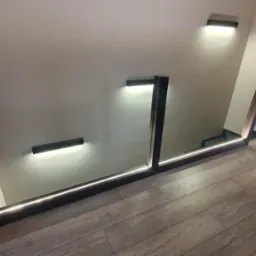 Nowoczesne schody z podświetlanymi stopniami i minimalistycznymi balustradami z ciemnego drewna, oświetlenie ścienne w kształcie pasków, widok z góry na drewnianą podłogę.