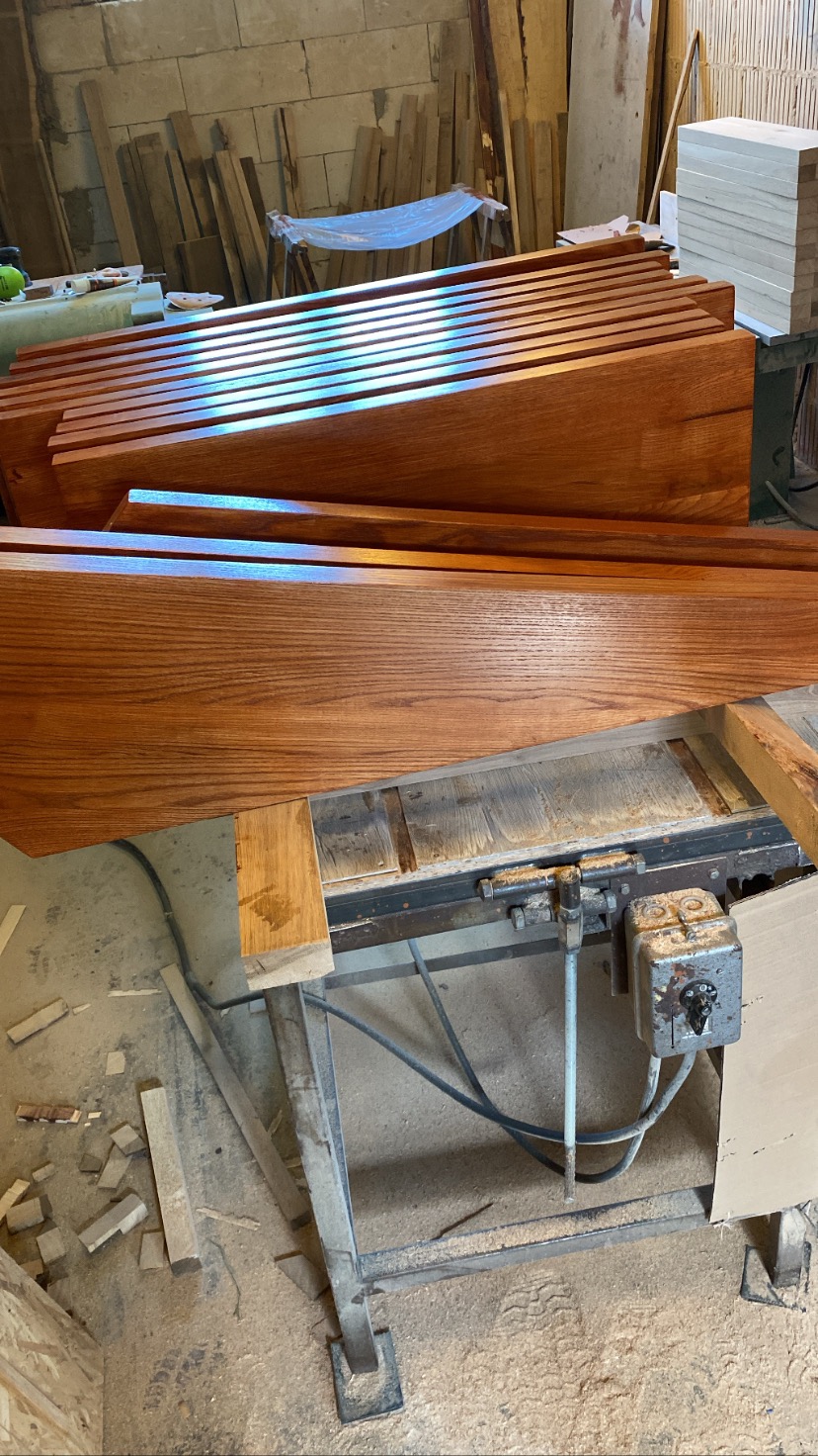 Drewniane elementy schodów, prawdopodobnie stopnie, ułożone w warsztacie na stole z piłą tarczową, widoczne ślady obróbki drewna.