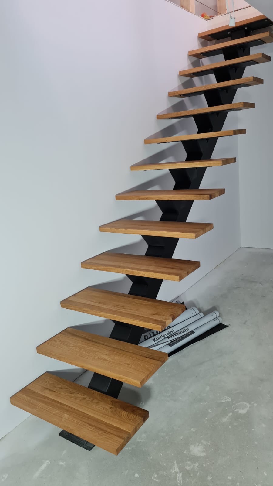 Nowoczesne schody wspornikowe z drewnianymi stopniami i czarną metalową konstrukcją nośną, widoczne materiały budowlane na podłodze, surowe wykończenie wnętrza.