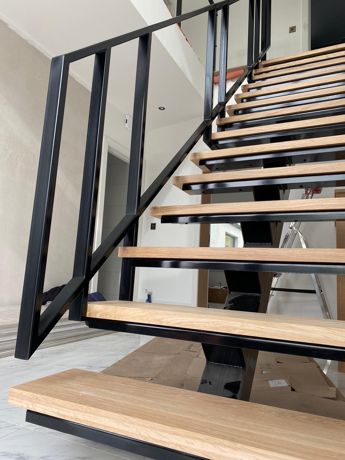 Nowoczesne schody z drewnianymi stopniami i czarną metalową konstrukcją, widoczne elementy montażowe i drabina w tle, sugerujące trwający montaż.