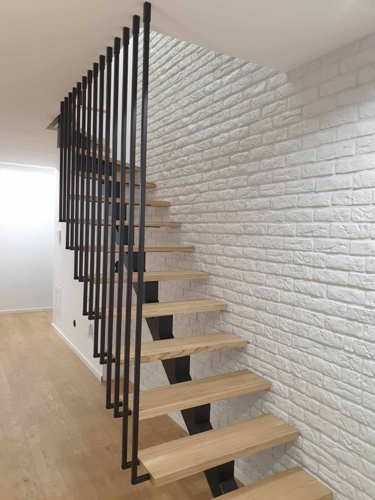 Nowoczesne schody z drewnianymi stopniami i czarną, minimalistyczną balustradą z pionowych prętów, na tle ściany z białej cegły.
