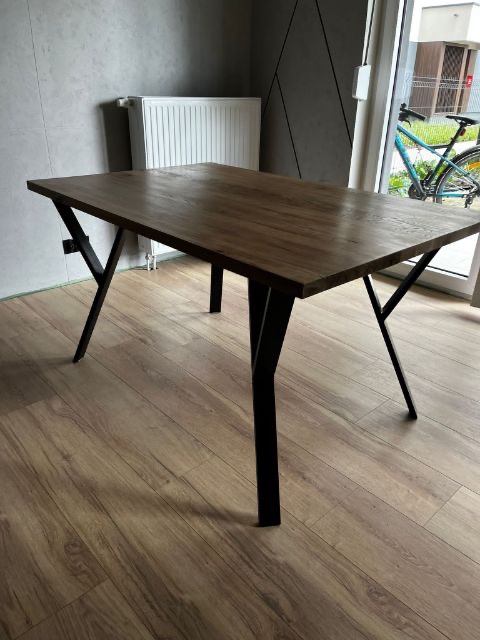 Stół z blatem z ciemnego drewna i czarnymi, metalowymi nogami w kształcie litery V, ustawiony w nowoczesnym wnętrzu z szarą ścianą i widocznym fragmentem roweru za oknem.