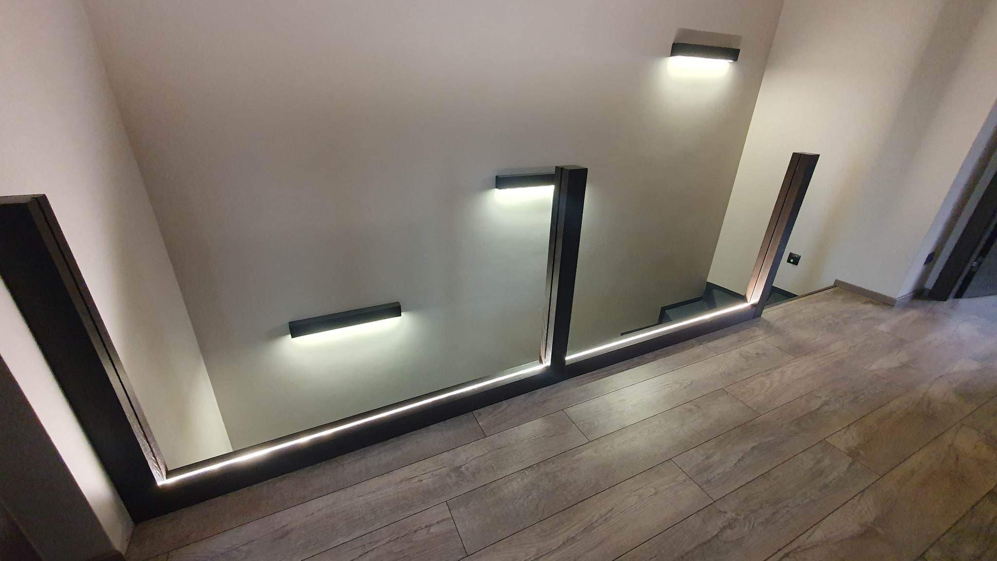 Nowoczesne schody z podświetlanymi stopniami i minimalistycznymi balustradami z ciemnego drewna, oświetlenie ścienne w kształcie pasków, widok z góry na drewnianą podłogę.