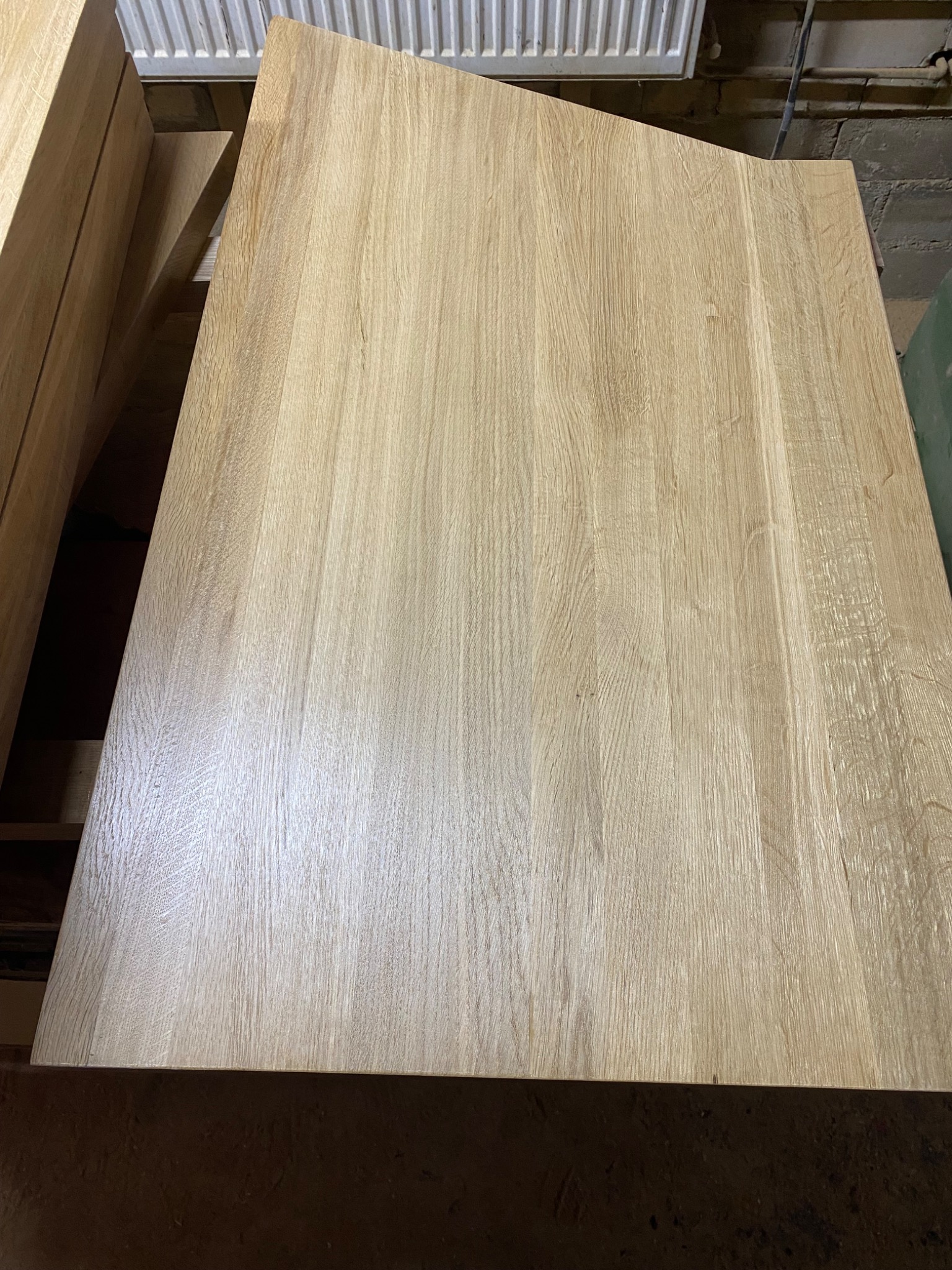 Jasny, dębowy blat o nieregularnym kształcie, widoczne słoje drewna, ułożony na konstrukcji w warsztacie, surowy stan wykończenia.