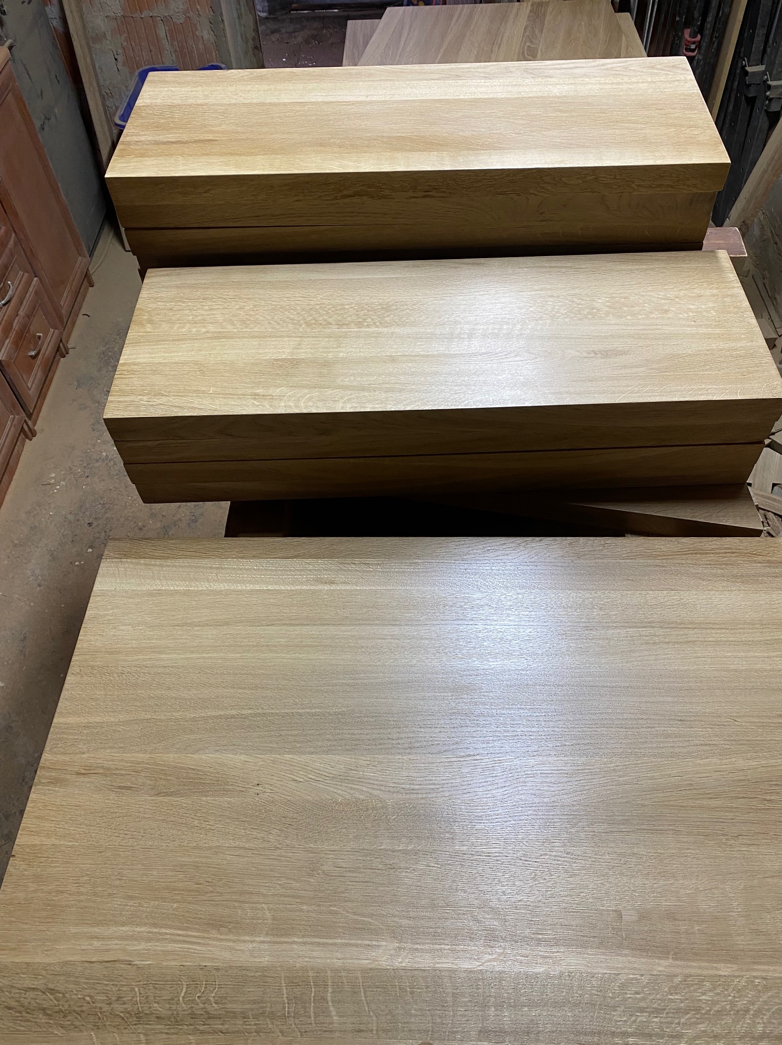 Stos wykończonych, jasnych blatów z drewna dębowego, ułożonych jeden na drugim w warsztacie stolarskim. Widoczne detale usłojenia drewna.