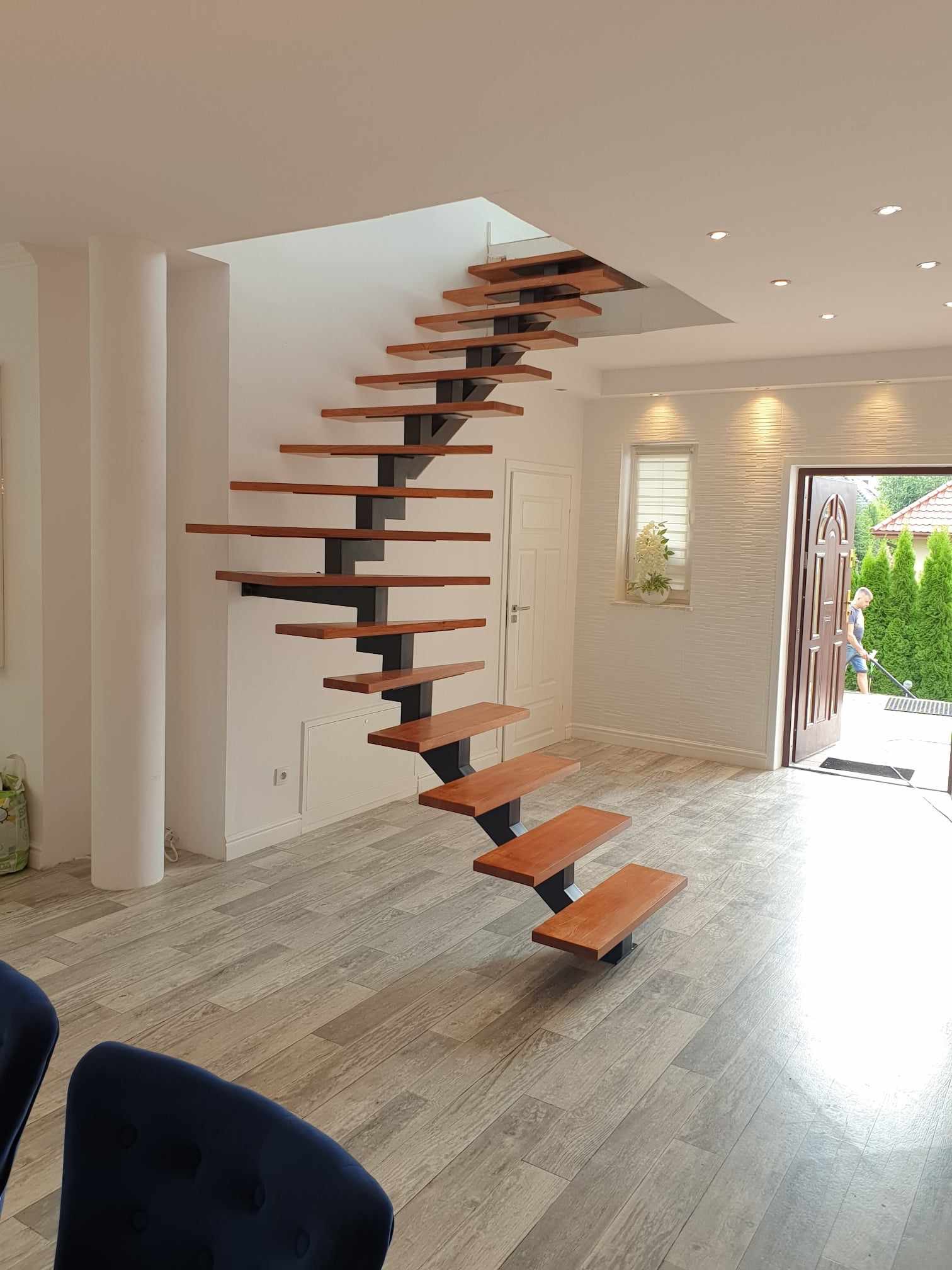 Nowoczesne schody wspornikowe z drewnianymi stopniami i metalową konstrukcją w jasnym wnętrzu domu, z widokiem na wejście i osobę koszącą trawę na zewnątrz.