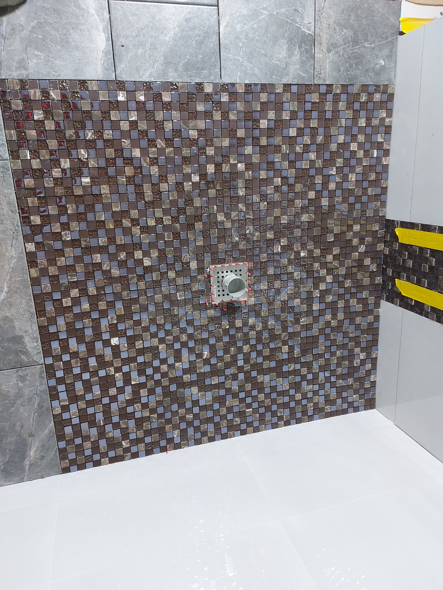 Fragment ściany łazienki wyłożonej mozaiką w odcieniach brązu i szarości, z widocznym odpływem liniowym zabezpieczonym czerwoną taśmą, oraz fragmentem szarej płytki imitującej marmur.