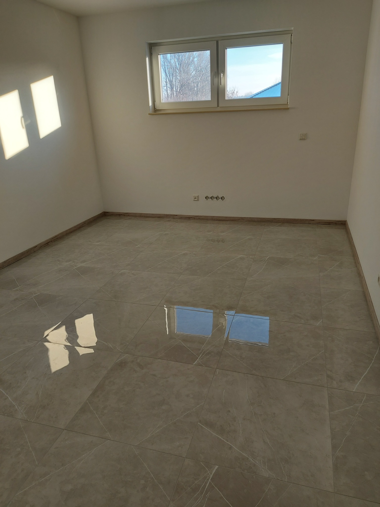 Puste pomieszczenie z beżowymi płytkami podłogowymi imitującymi marmur, białe ściany, okno z widokiem na drzewa, wpadające promienie słoneczne na ścianie i odbicie okna w podłodze.