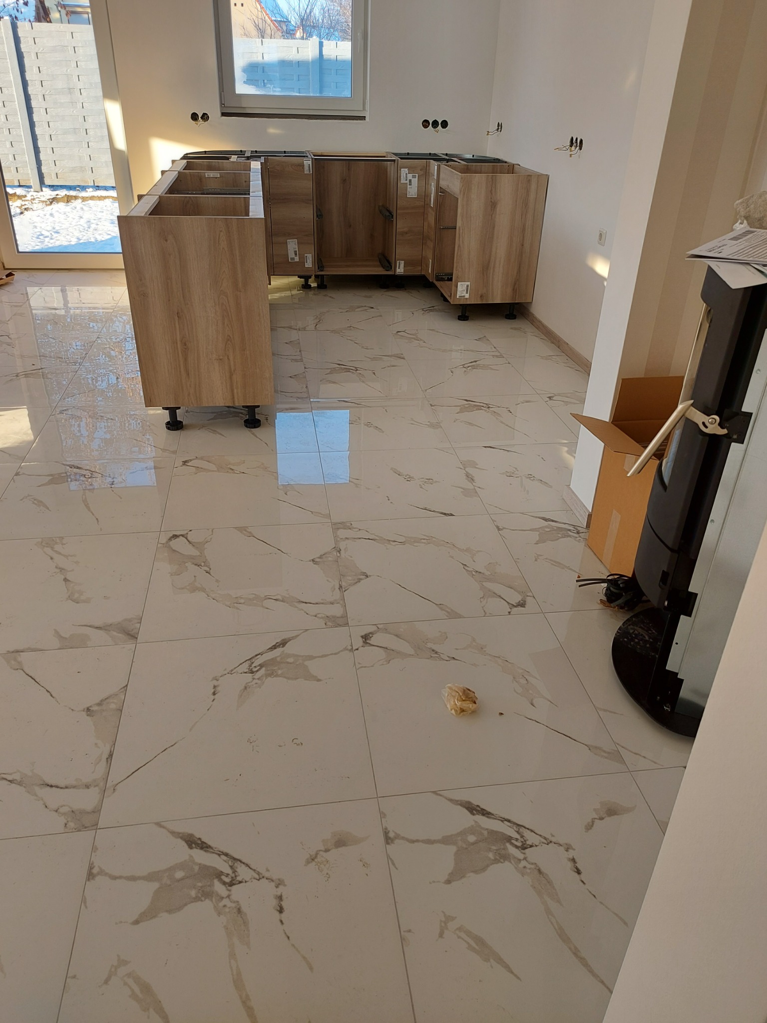 Częściowo zamontowane, drewnopodobne szafki kuchenne na regulowanych nóżkach, na podłodze z białych płytek imitujących marmur, widoczne przygotowane instalacje elektryczne na ścianie.