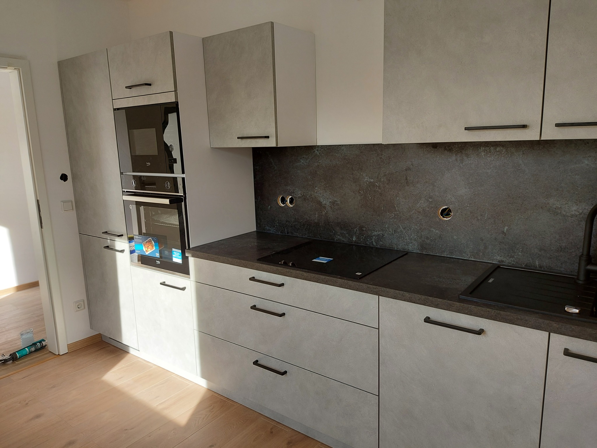 Nowoczesna kuchnia z szarymi, matowymi frontami szafek, czarnym blatem i sprzętem AGD; widoczna płyta indukcyjna i piekarnik w zabudowie, na podłodze panele drewnopodobne.