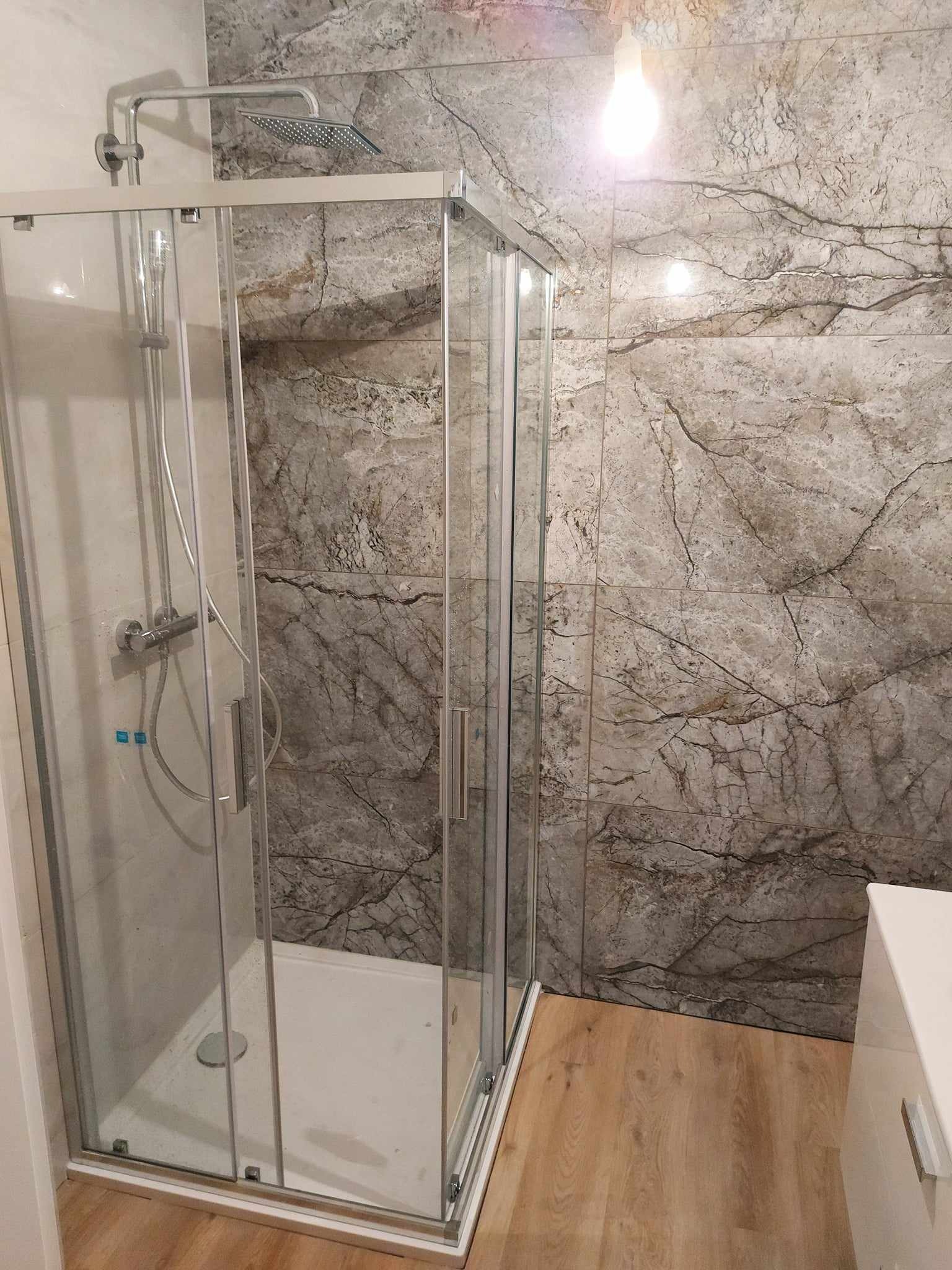 Szklana kabina prysznicowa z przesuwnymi drzwiami, zamontowana na białym brodziku, z szarymi płytkami imitującymi marmur na ścianie i panelami podłogowymi