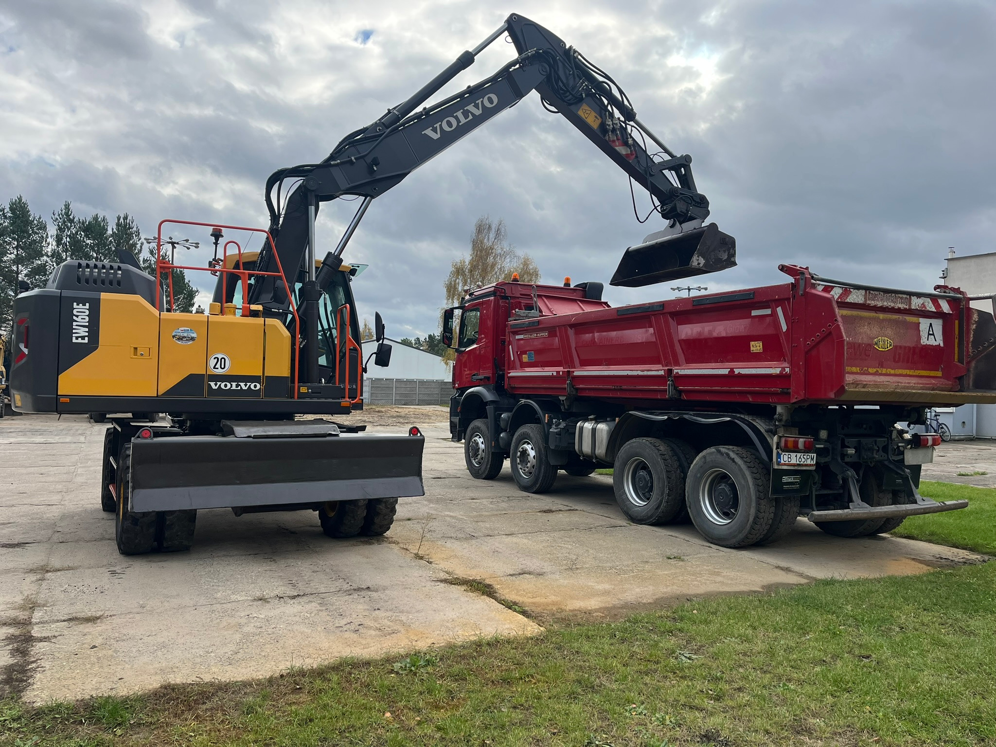 Żółto-czarna koparka Volvo EW160E na kołach, nabiera ziemię do czerwonej ciężarówki. Praca w toku, pochmurne niebo w tle. Widoczne detale maszyn.