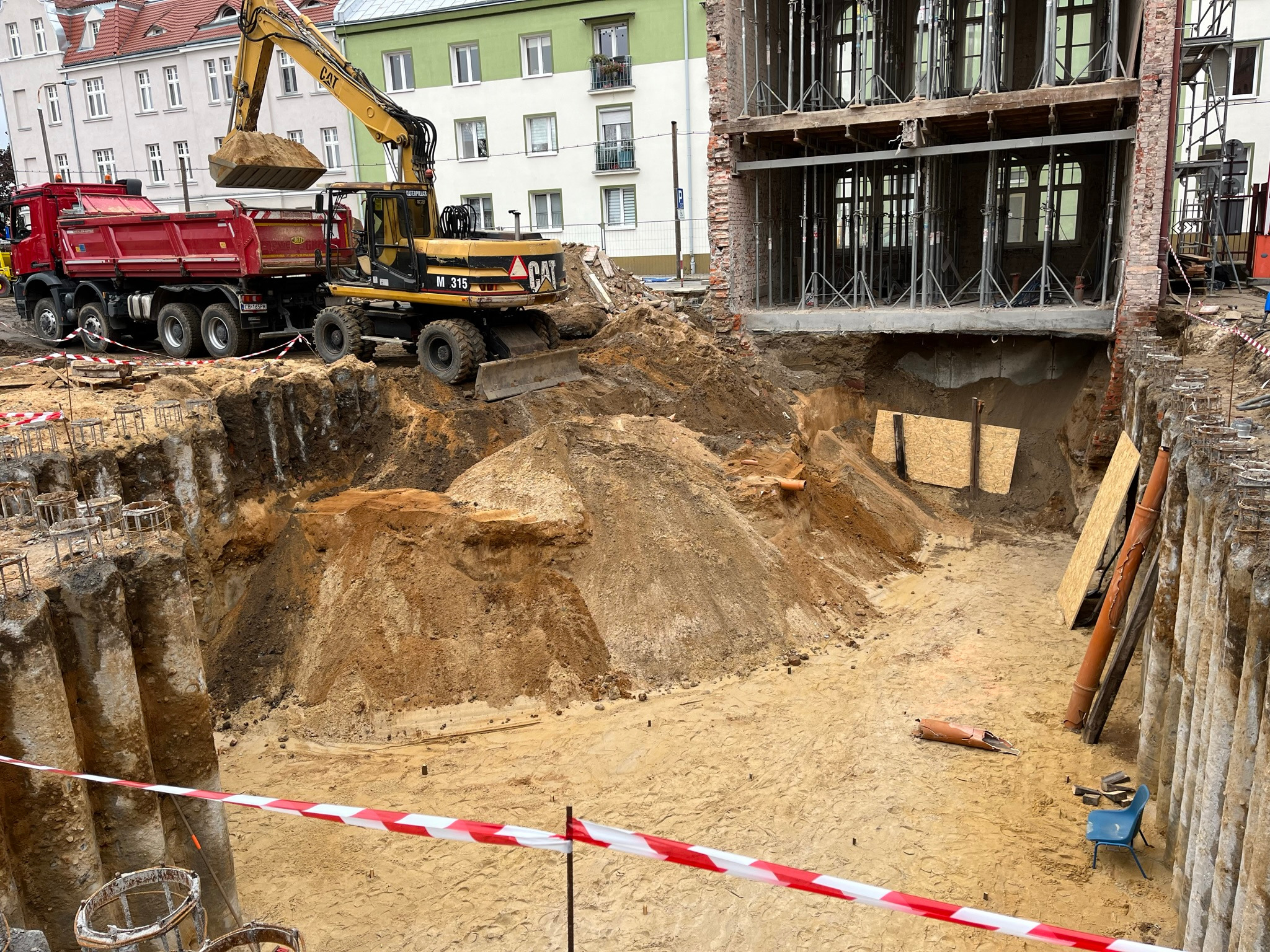 Żółta koparka CAT M315 ładuje ziemię do czerwonej ciężarówki na placu budowy z odsłoniętymi fundamentami budynku w tle.