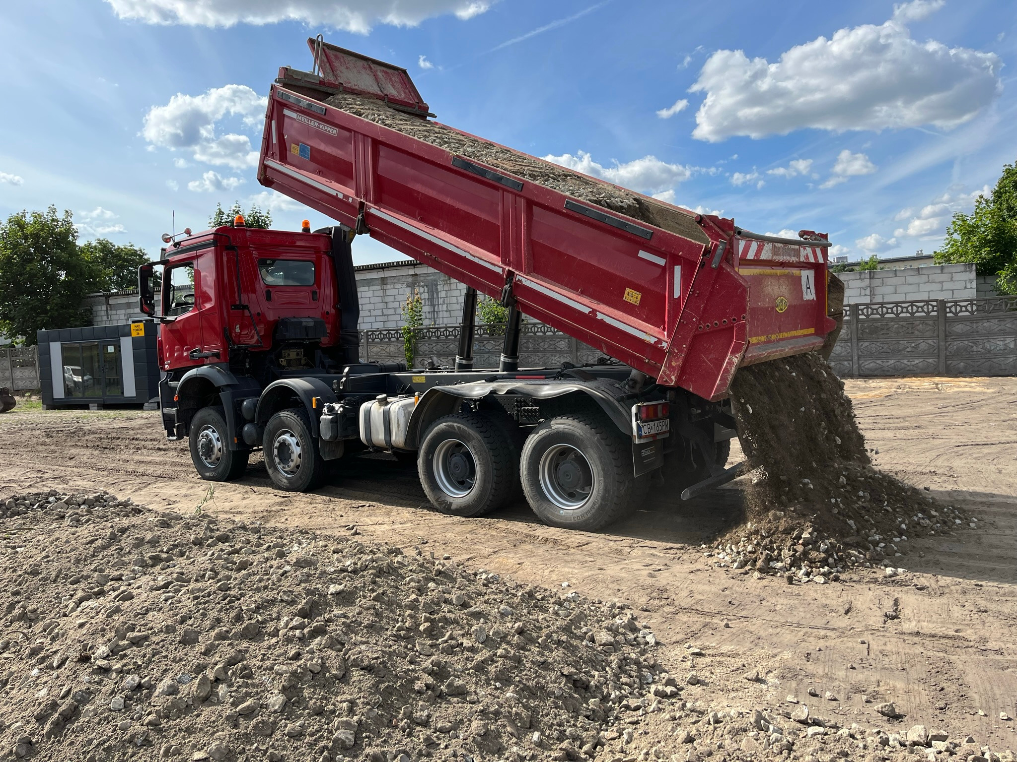 Czerwona wywrotka z podniesioną skrzynią ładunkową wysypuje żwir na ziemię, widoczne ogrodzenie z białej cegły w tle i błękitne niebo z chmurami.