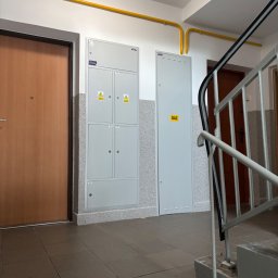 Łukasz Brzegowy - Szara, wielosekcyjna rozdzielnica elektryczna z naklejkami ostrzegawczymi i szafka gazowa z napisem GAZ na ścianie korytarza, z żółtymi rurami gazowymi.