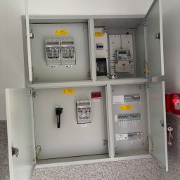 Łukasz Brzegowy - Dwie otwarte, szare rozdzielnice elektryczne z licznikiem, głównym wyłącznikiem i szeregiem zabezpieczeń, zamontowane na ścianie nad nakrapianą podłogą, obok czerwonego przycisku alarmowego.