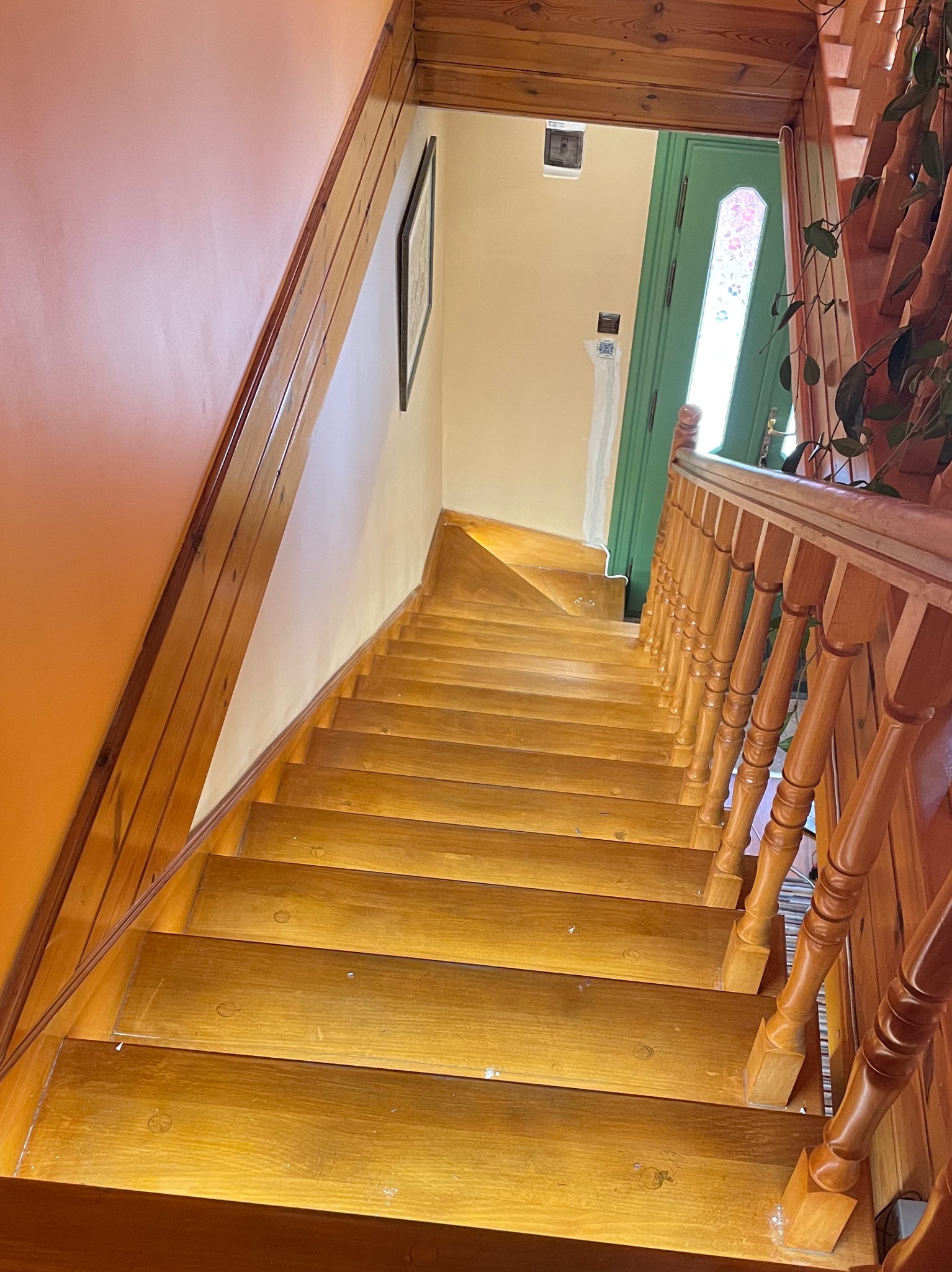 Drewniane schody z poręczą, widok z góry, ściana pomalowana na kolor brzoskwiniowy, na ścianie wisi obrazek, zielone drzwi w tle, skrzynka elektryczna na ścianie przy drzwiach.